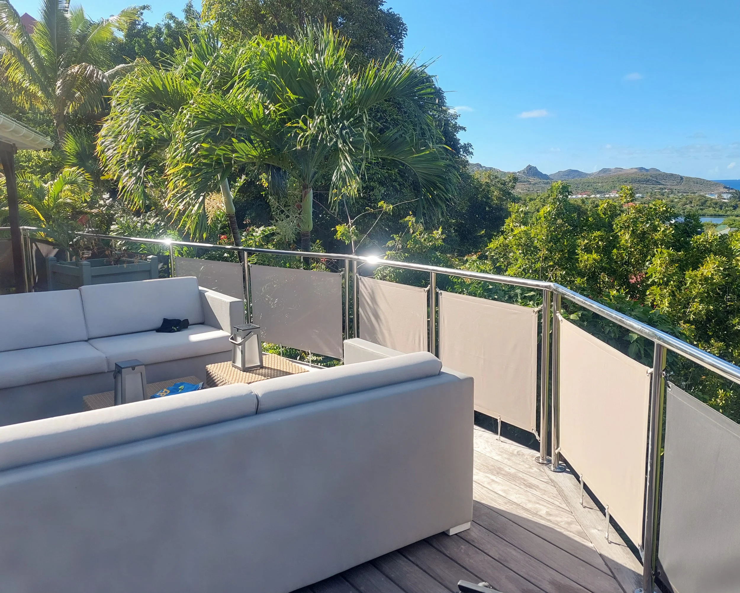 A balcony with white outdoor couches and coffee tables, overlooking green trees, hills, and a clear blue sky on a sunny day.