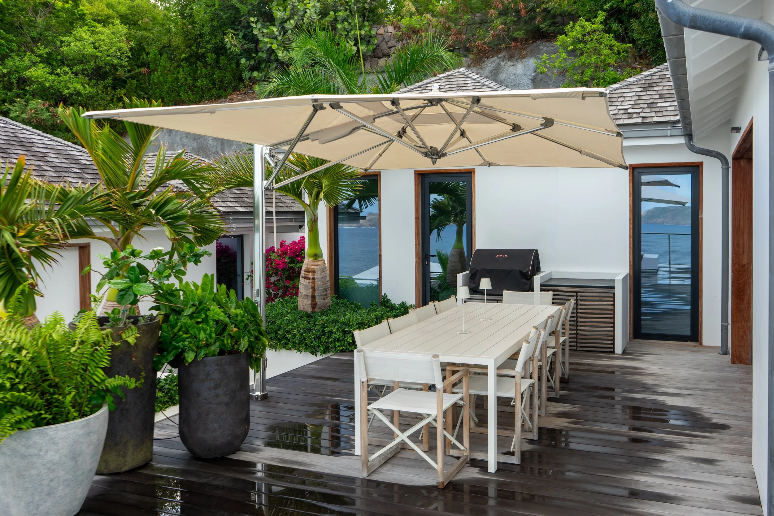 A patio area with a large cream-colored umbrella over a white outdoor dining table and chairs, surrounded by potted plants and greenery, with a view of the ocean in the background.