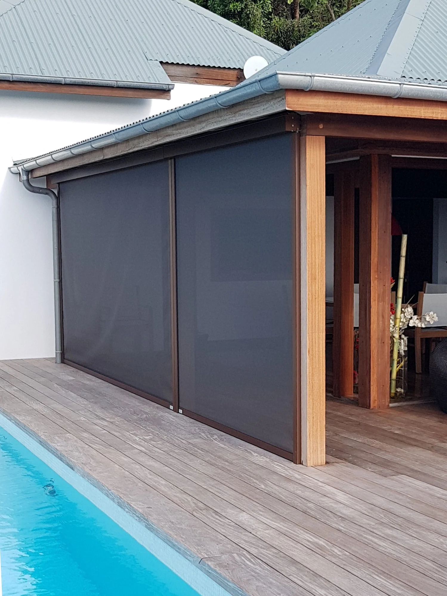 Backyard swimming pool with a wooden deck, gray pool cover, and a pool house with a corrugated metal roof and wooden framing.