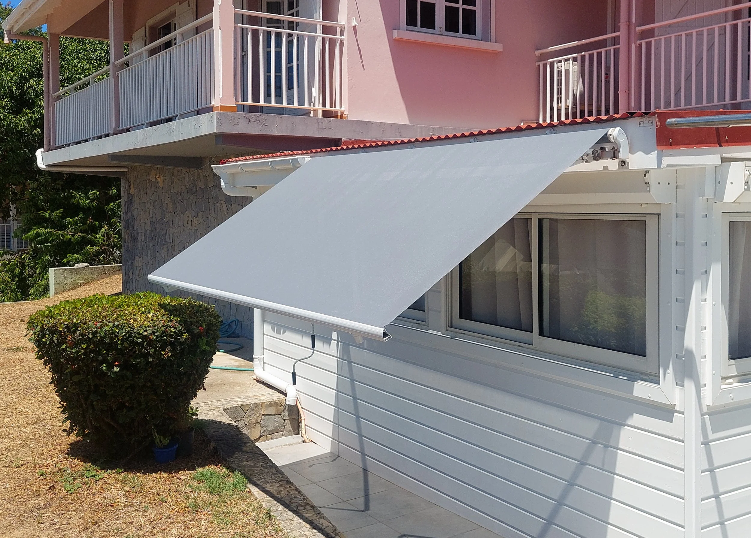 The image shows the exterior of a two-story house with pink walls and white balconies with railings. There is an awning with a retractable white shade extending from the side of the house, above a patio area. A bush is in the foreground on a dirt pat
