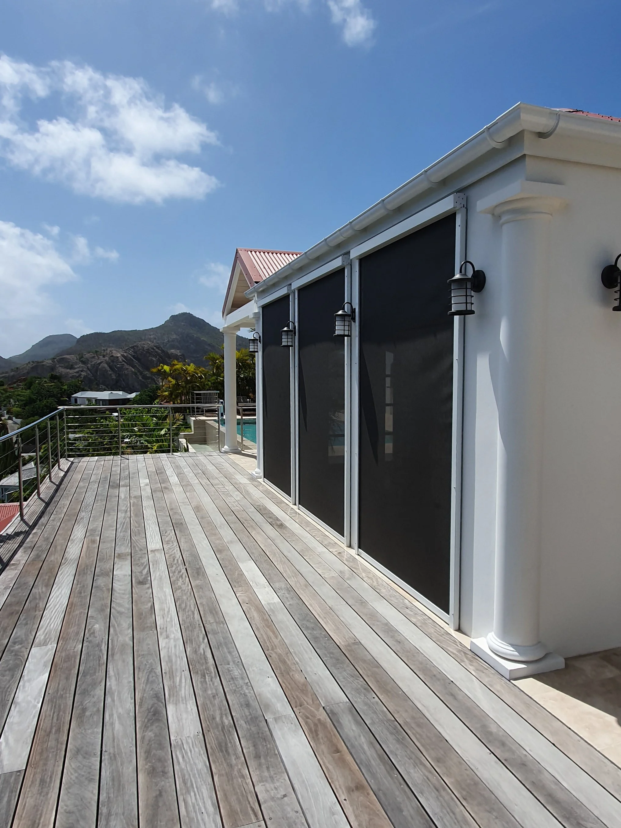 A spacious outdoor deck with wooden planks, white columns, black window screens, and a view of mountains and blue sky with clouds in the background.