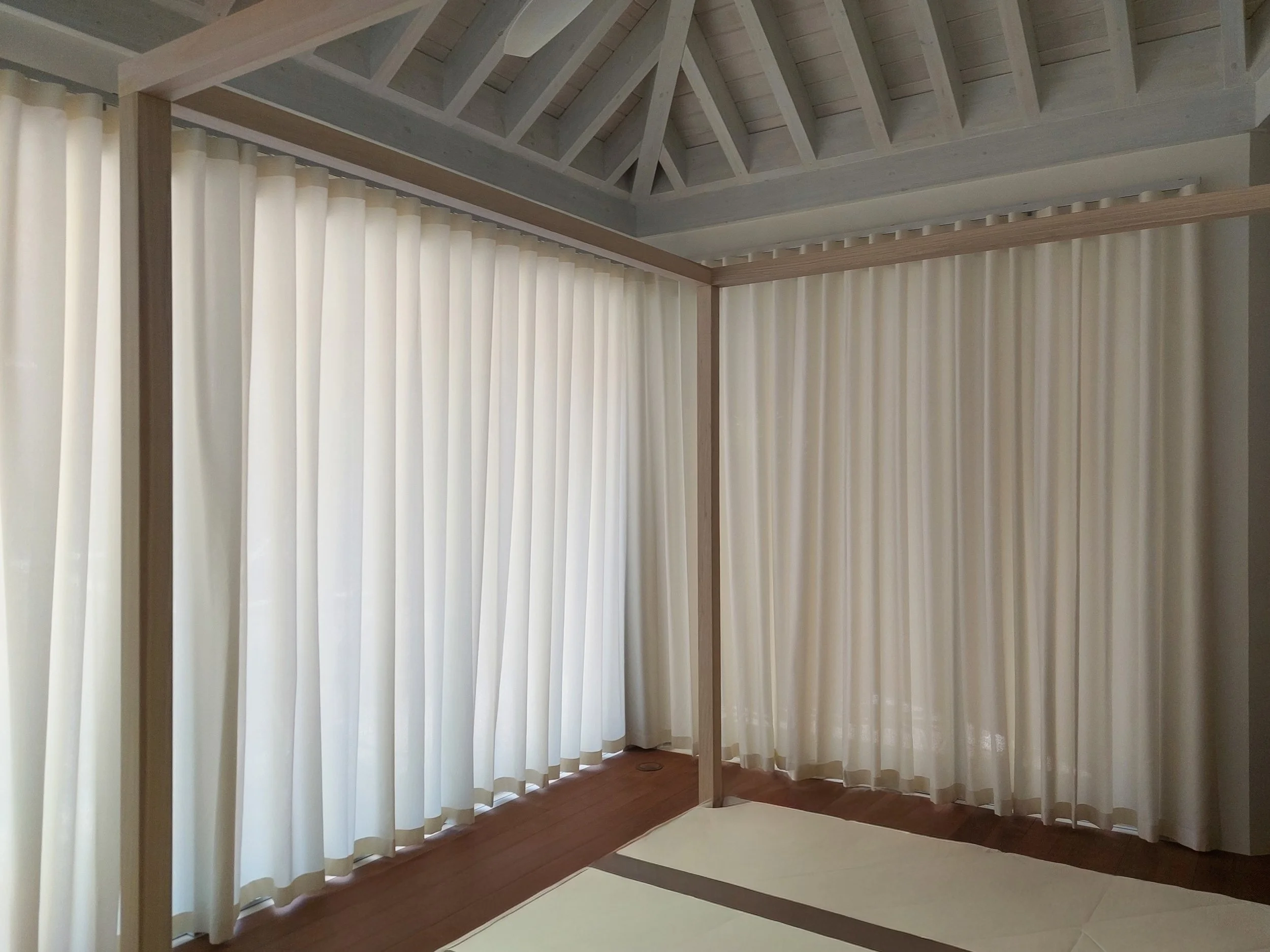 Interior of a room with large white curtains hanging from ceiling to floor on three sides, with a wooden frame structure supporting the ceiling and wall.