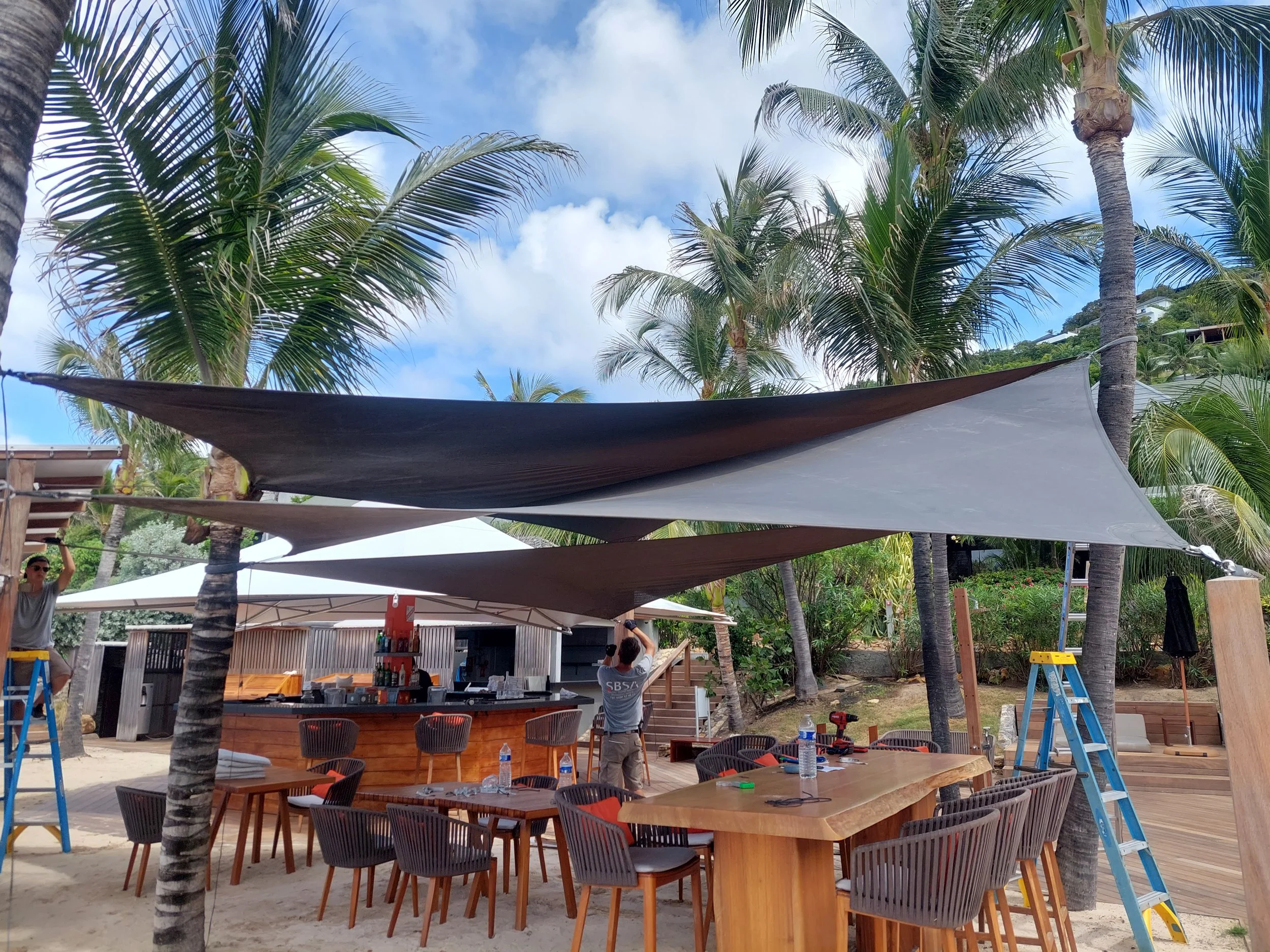 Setting up an outdoor event under palm trees with tables, chairs, and shade sails on a sandy beach.