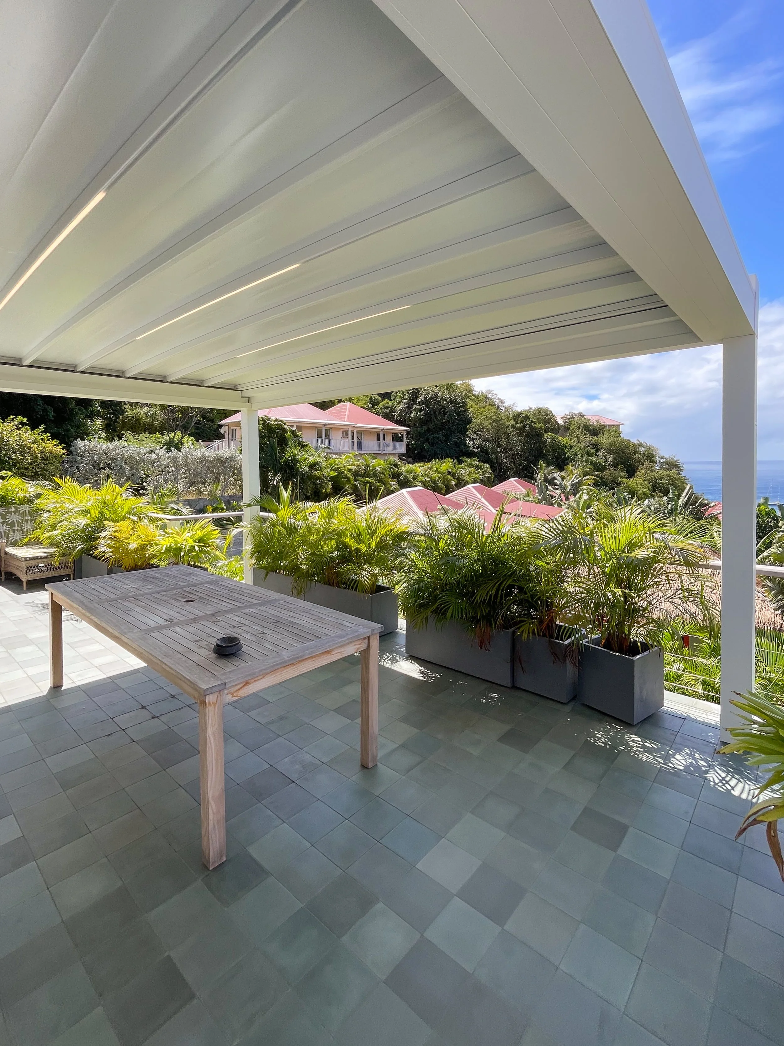 A covered outdoor patio area with a wooden table and green potted plants, overlooking houses and trees with a scenic view of the ocean in the distance under a partly cloudy sky.