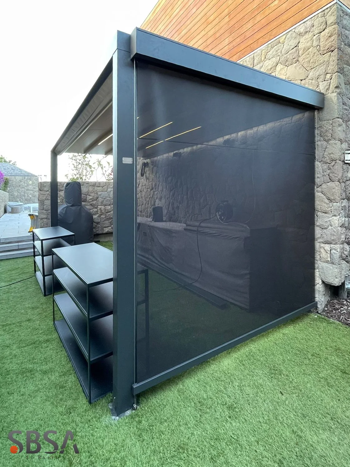 An outdoor covered patio area with a black metal frame, black shelving units, and a stone and wood exterior wall. The patio features a black tinted screen or wall on one side, with grass on the ground.