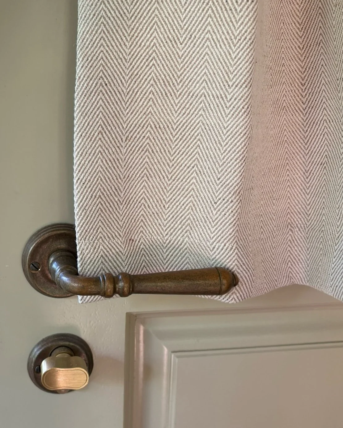 Close-up of a beige curtain with a herringbone pattern hanging in front of a door, with a wooden door handle and a lock visible.