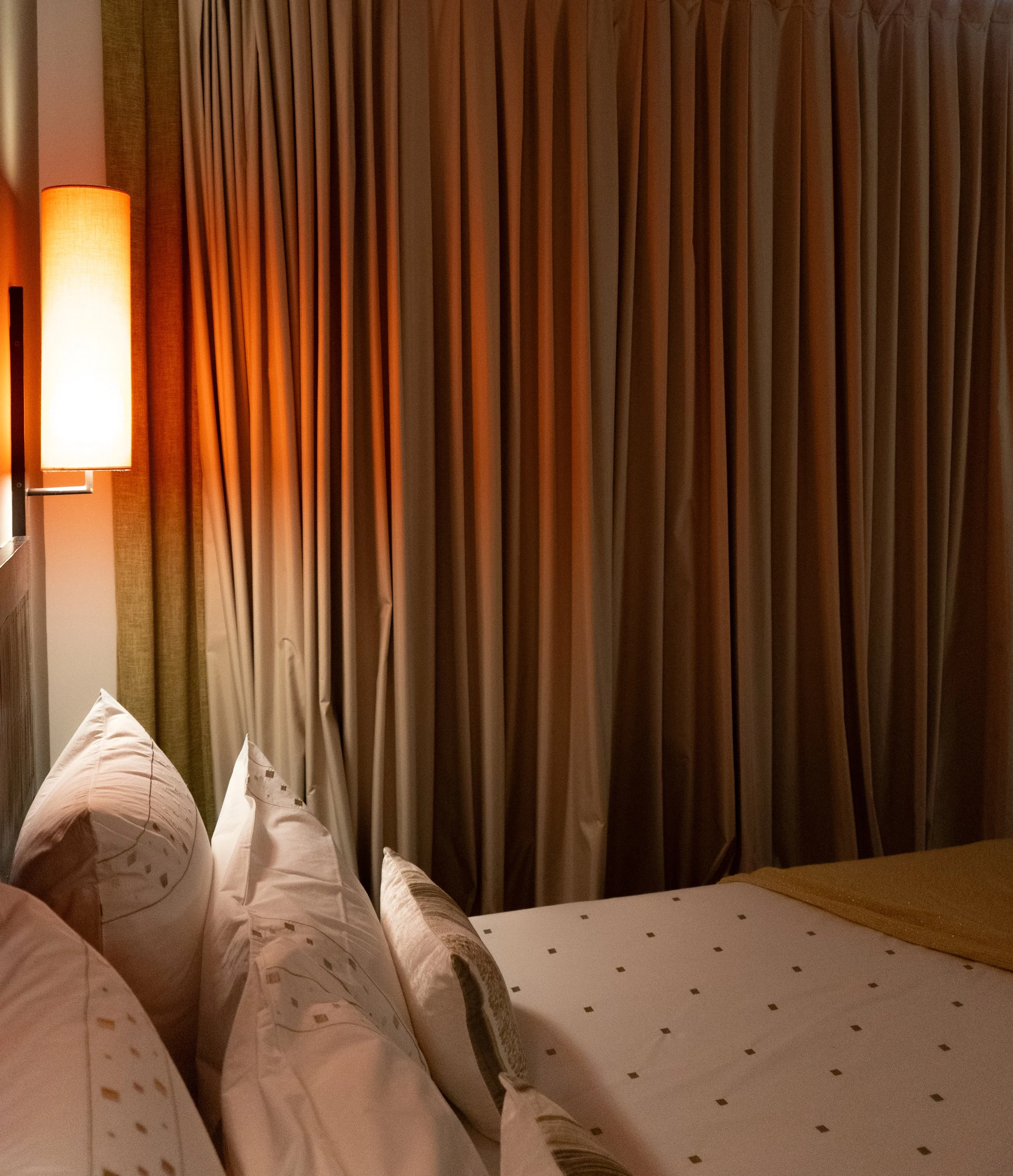 Une chambre d’hôtel avec des rideaux beiges, un lit garni d’oreillers et une lampe de chevet allumée diffusant une lumière chaleureuse.