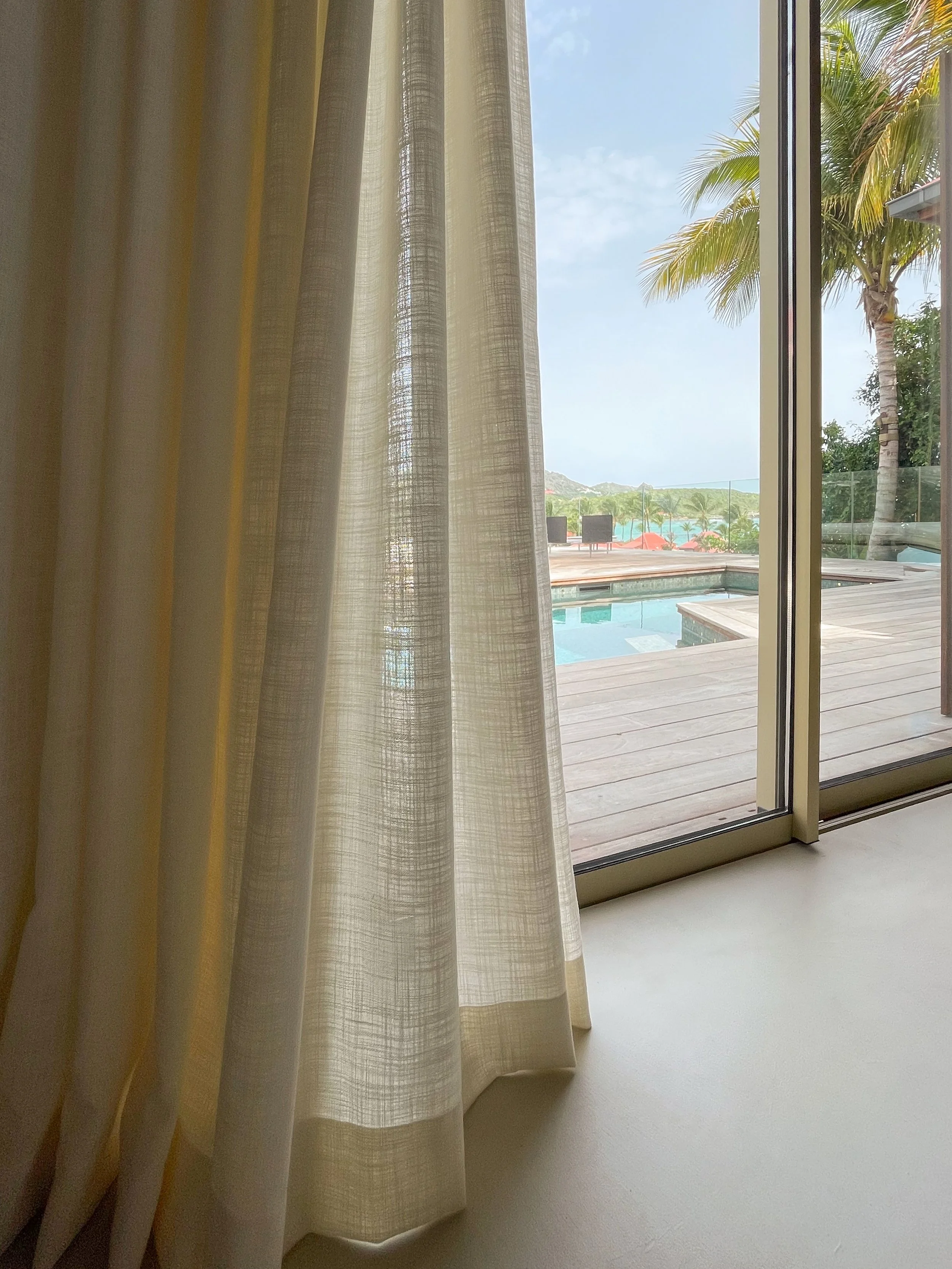 Une vue depuis un intérieur d'une pièce avec des rideaux légers de couleur beige ou crème, donnant sur une terrasse en bois avec une piscine. En arrière-plan, on aperçoit des palmiers et une mer ou un océan sous un ciel partiellement nuageux.