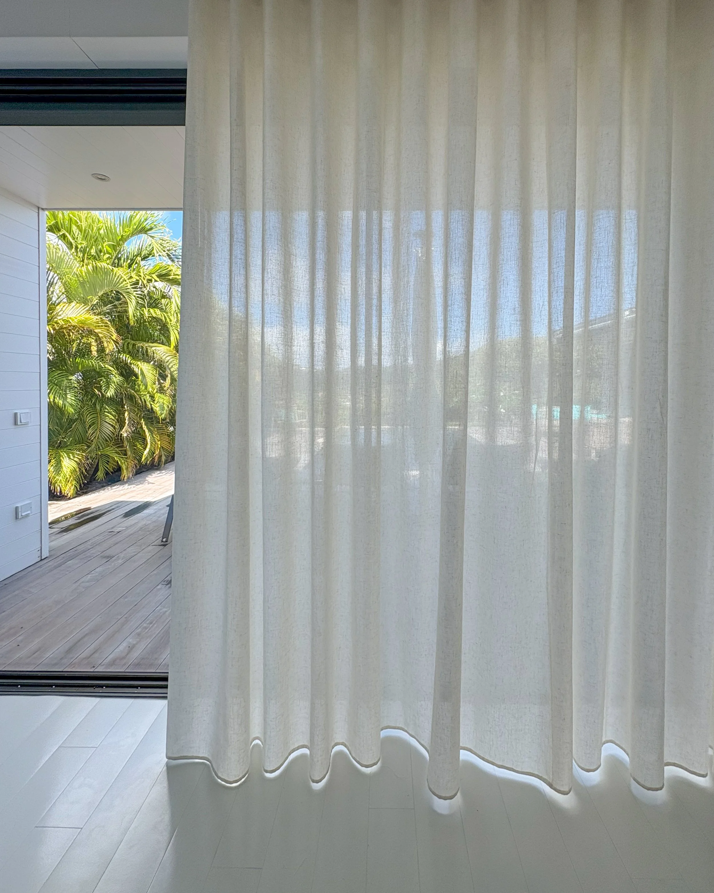 White curtains hanging on a window with sunlight filtering through, overlooking a garden with leafy green plants outside.