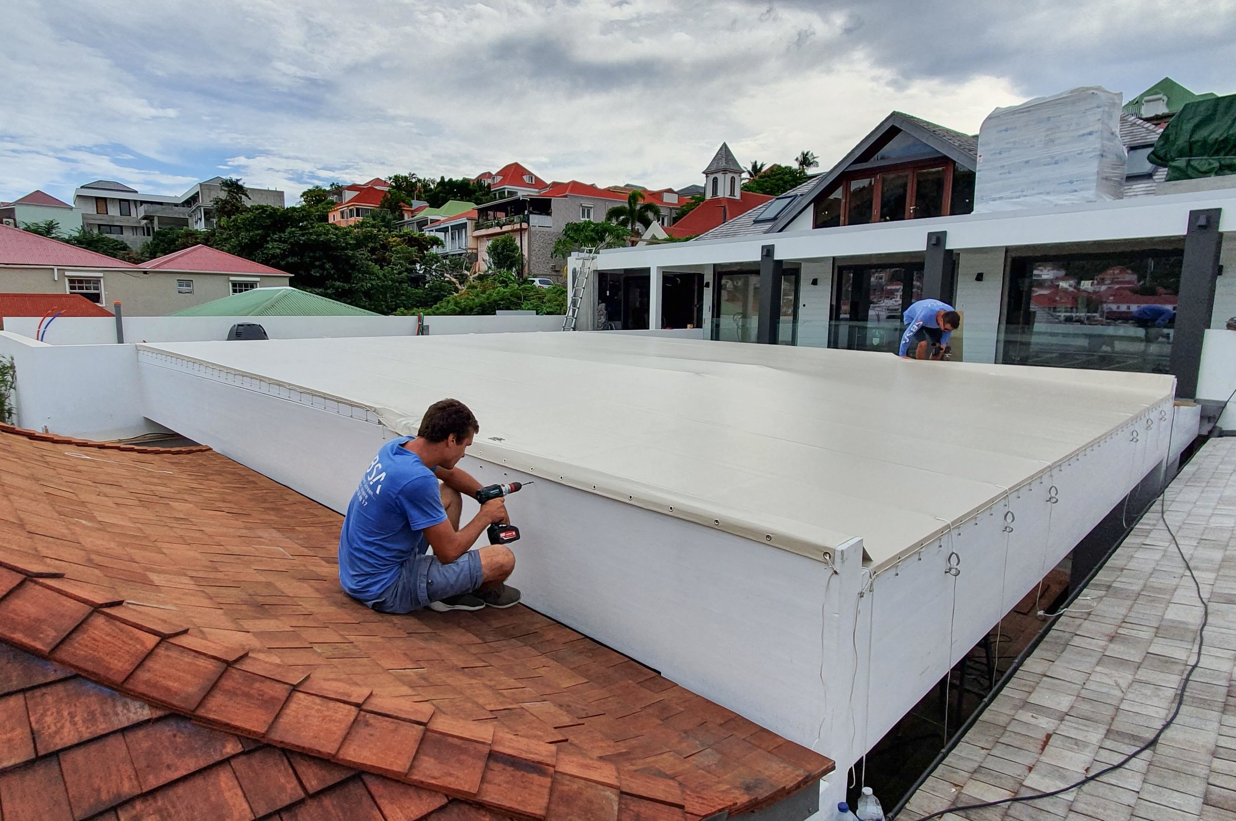 Two workers installing a white awning or shade on a house's balcony, with a background of residential houses and a cloudy sky.