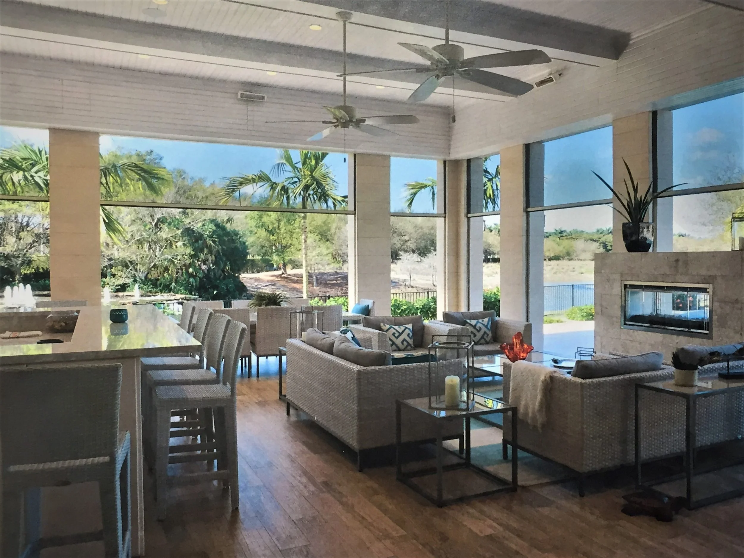 A spacious indoor living area with a sloped ceiling, ceiling fans, large windows, and patio doors overlooking a garden with trees. The room is furnished with a dining table, chairs, sofas, and decorative pillows.