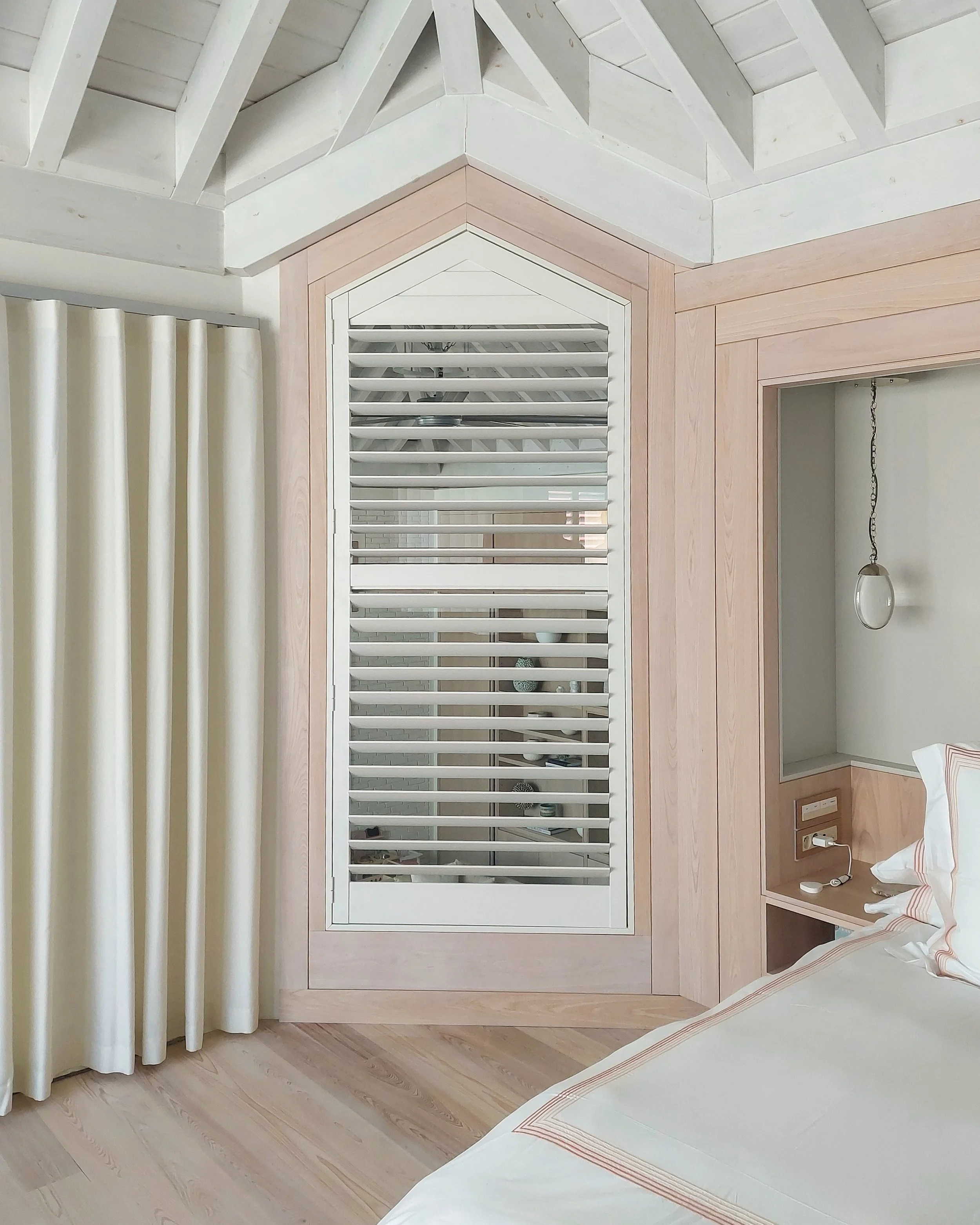 Une chambre avec un parquet en bois clair, des rideaux beiges et un lit partiellement visible dans le coin inférieur droit. La pièce est dotée d’un habillage mural en bois comprenant une fenêtre ou une grille d’aération blanche à lames horizontales, 