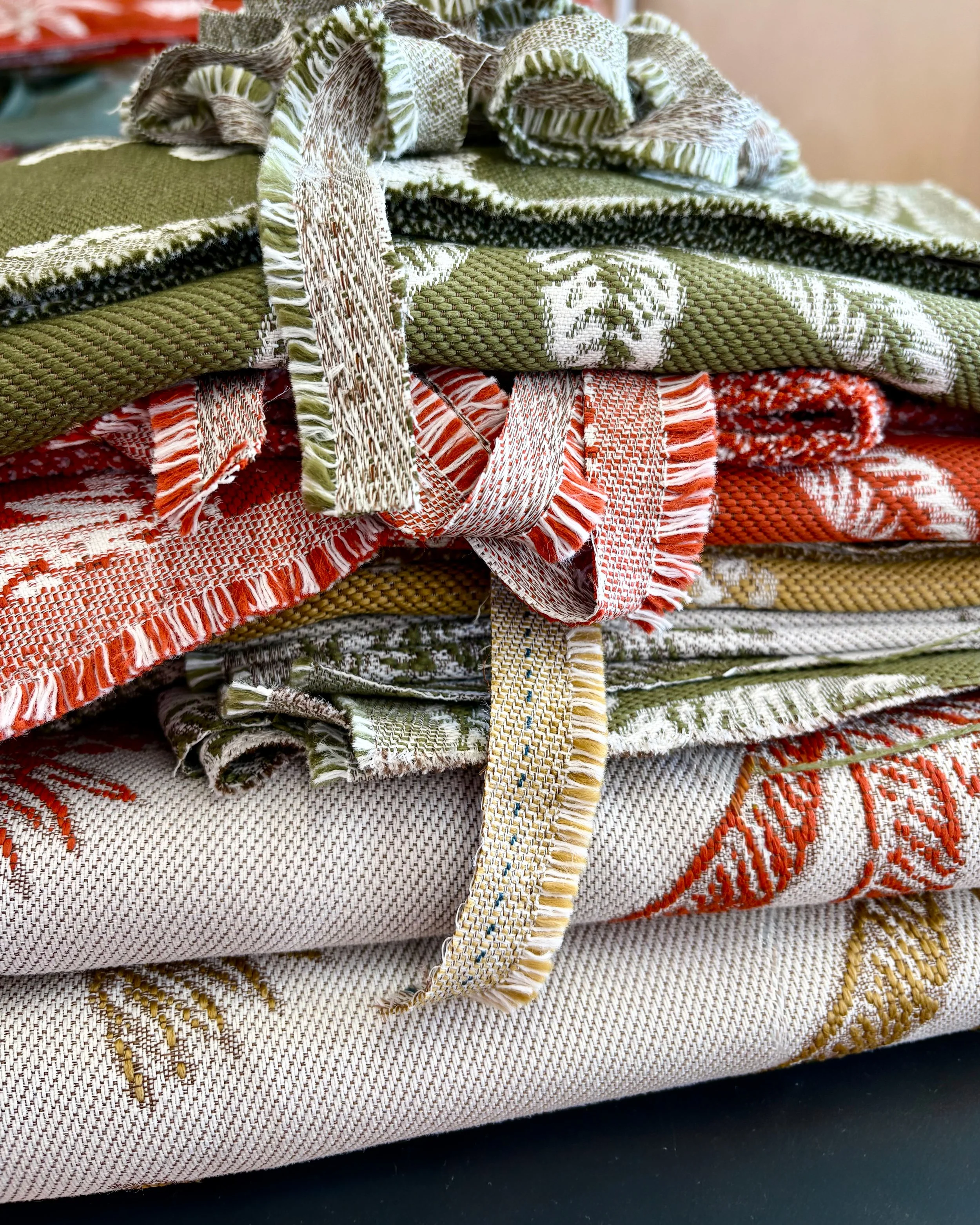 Stack of folded textiles with various woven patterns and fringed edges in green, orange, beige, and white colors.