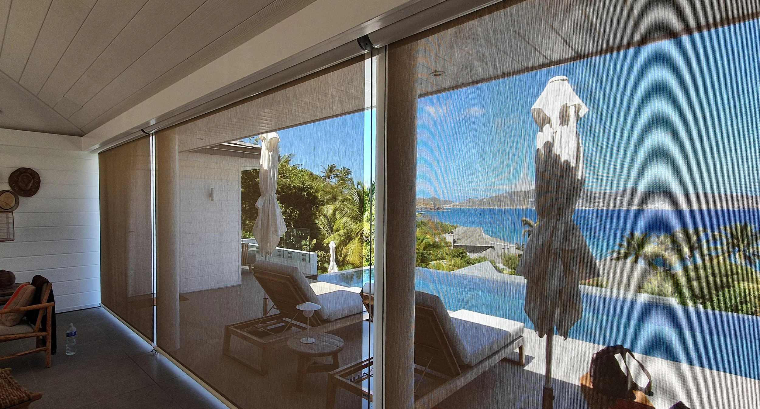 Interior view of a modern living room with electric vertical stores opening to a balcony with umbrellas, outdoor seating, palm trees, and a scenic ocean view with mountains in the distance under a clear blue sky.