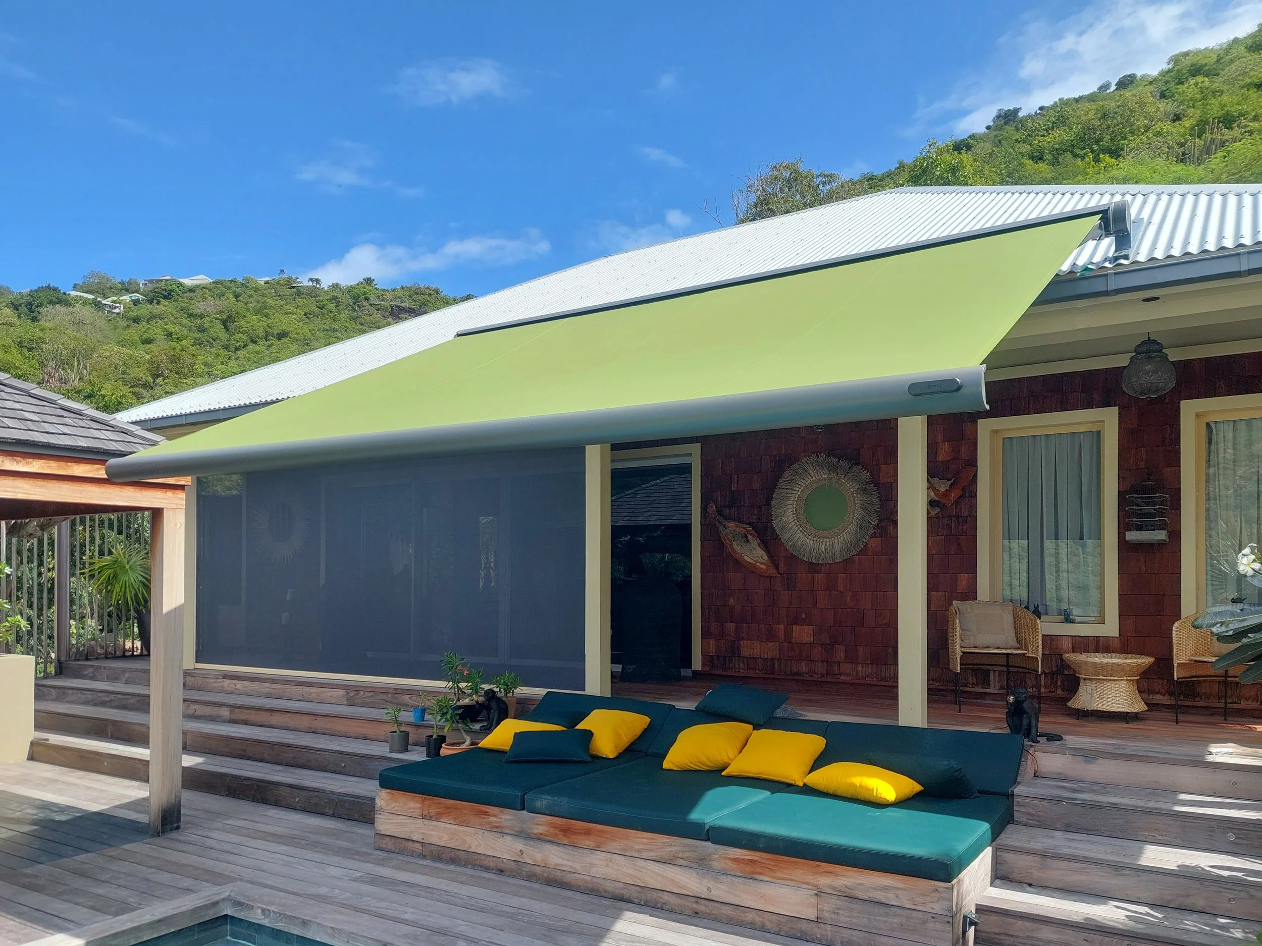 A backyard patio with wooden decking, a shade sail, and a seating area with teal cushions and yellow pillows, overlooking a house with red brick walls and a mountain with lush greenery in the background.