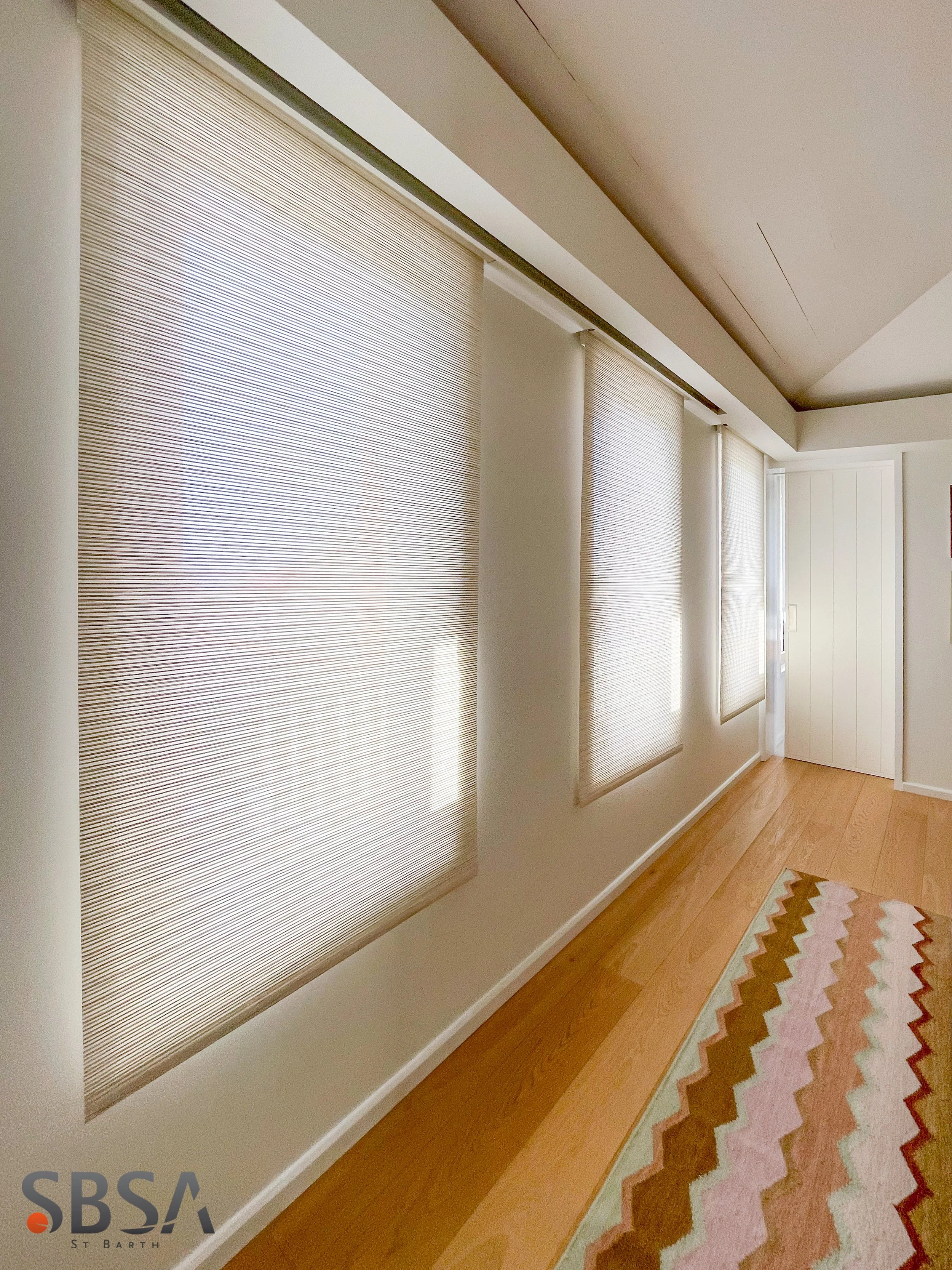 Multiple window blinds covering large windows in a room, next to a colorful zigzag-patterned rug on hardwood floor.
