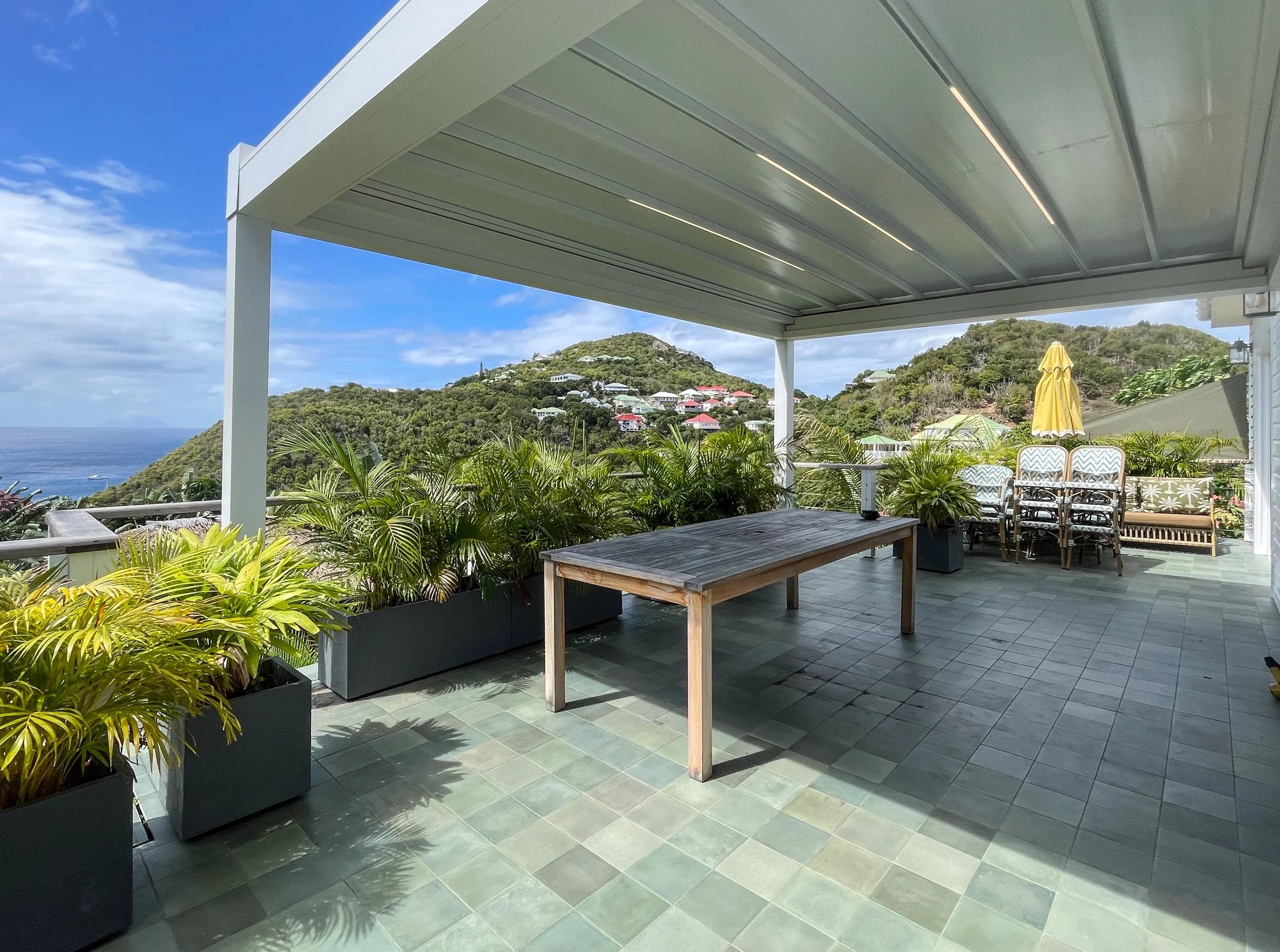 Covered outdoor patio area with potted plants, a wooden table, stacked chairs, and a view of lush green hills and colorful houses in the background.