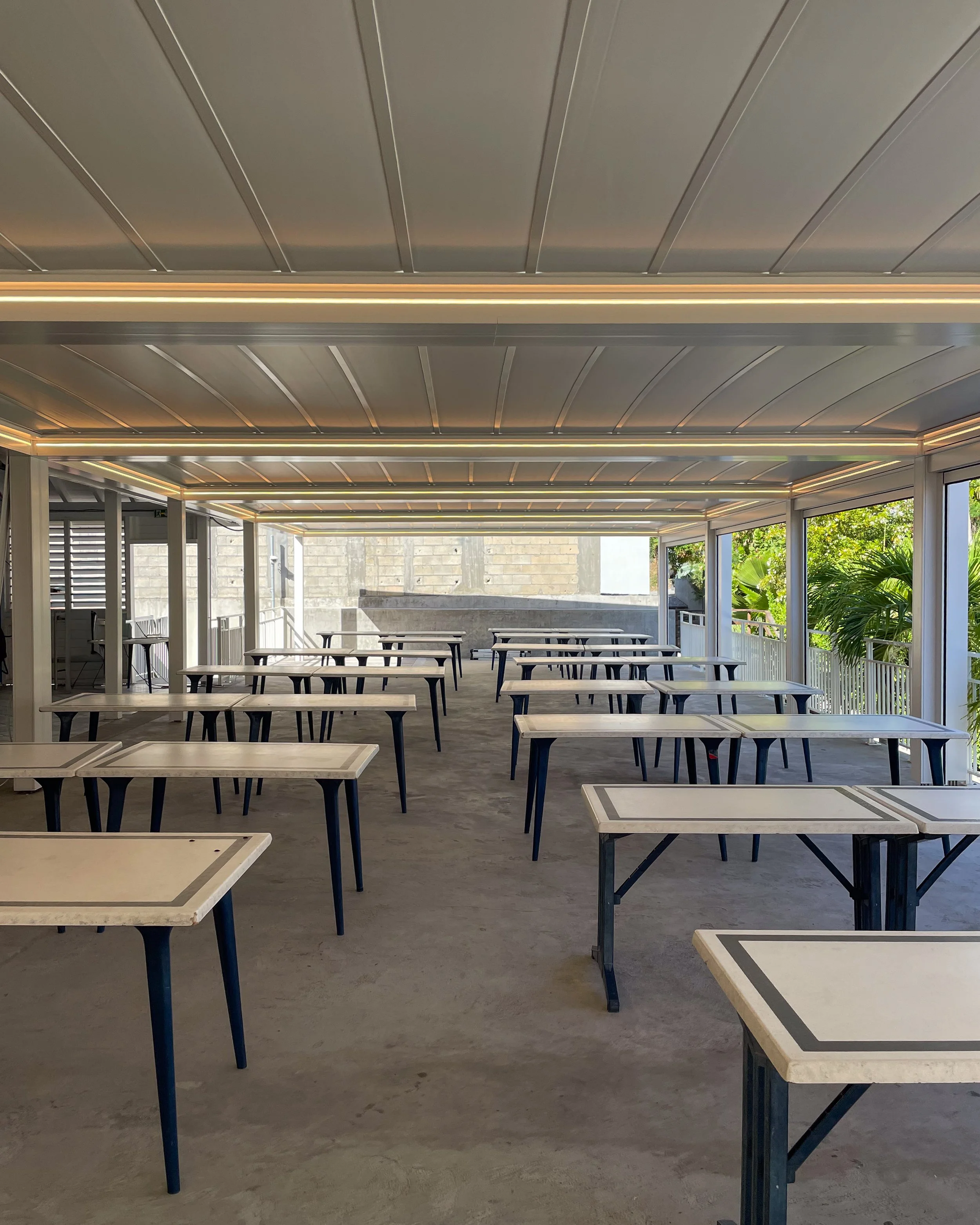 Indoor space with multiple tables and chairs, large windows on the right side, exposed ceiling with recessed lighting, and greenery outside.