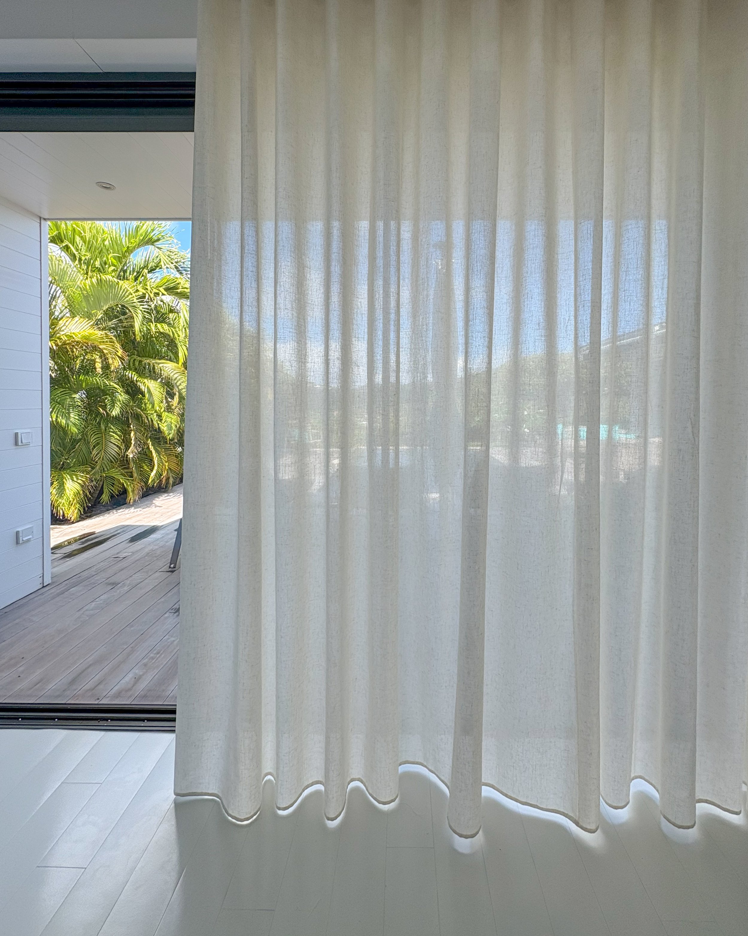 Vue intérieure d’une pièce avec un rideau blanc transparent suspendu devant une grande fenêtre. À l’extérieur, on aperçoit des palmiers verts et un ciel bleu. La pièce est dotée d’un sol carrelé blanc, et une partie d’un patio extérieur est visible.