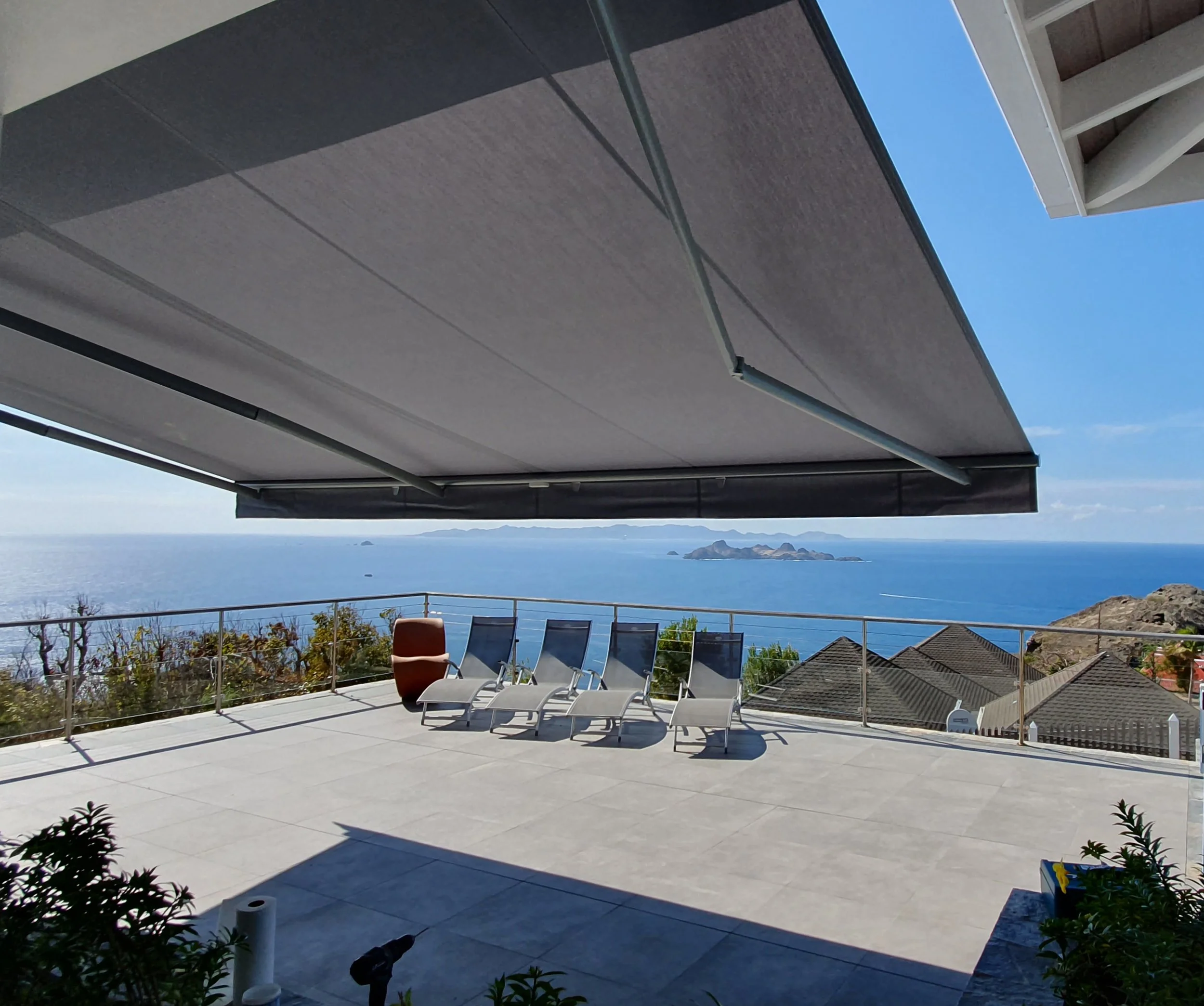 View of a spacious balcony with lounge chairs, overlooking the ocean and distant islands, shaded by an adjustable awning.