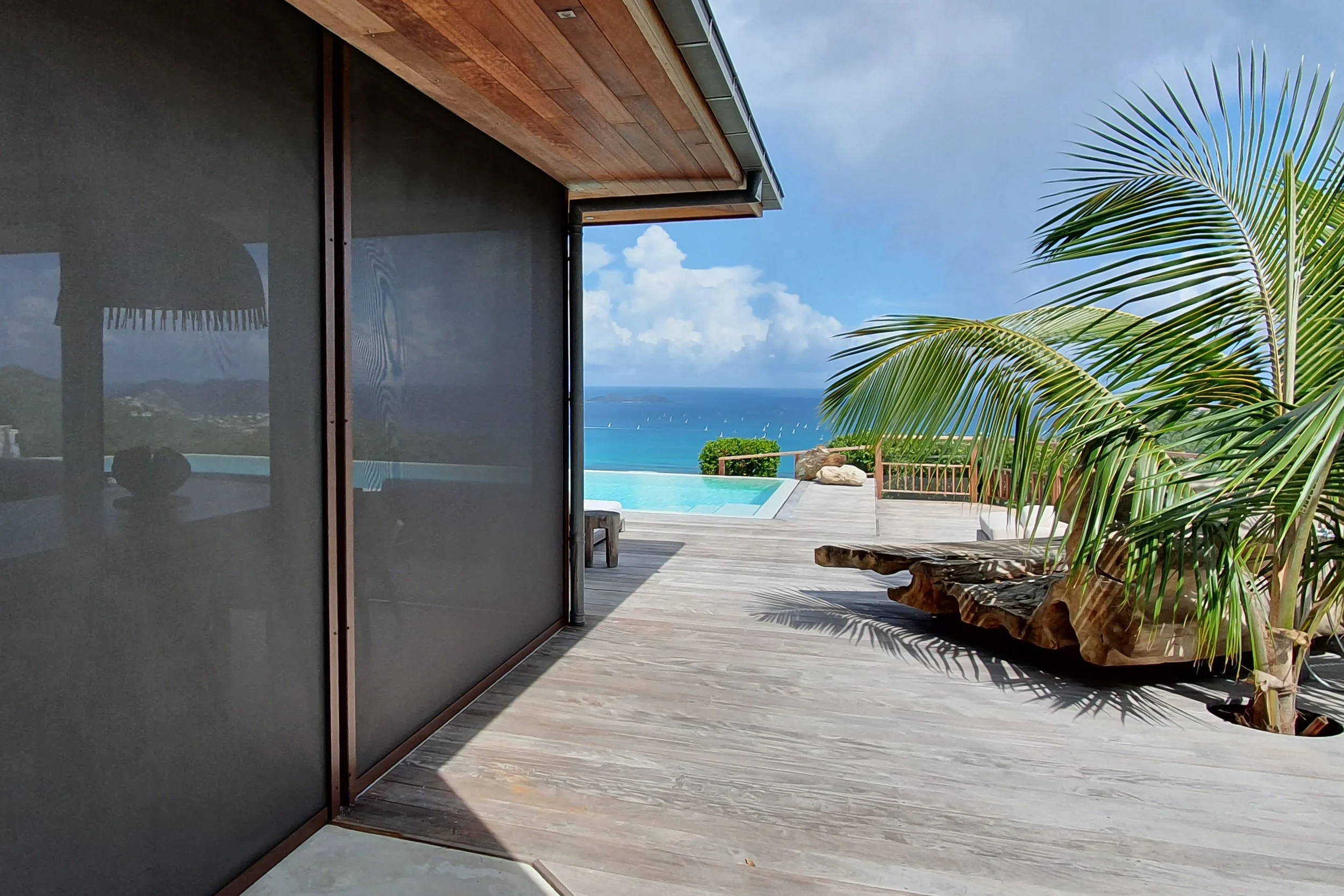 View from a patio showing a wooden deck, a modern outdoor bench made from a large curved piece of wood, a palm tree, and an infinity pool overlooking the ocean with several sailboats.
