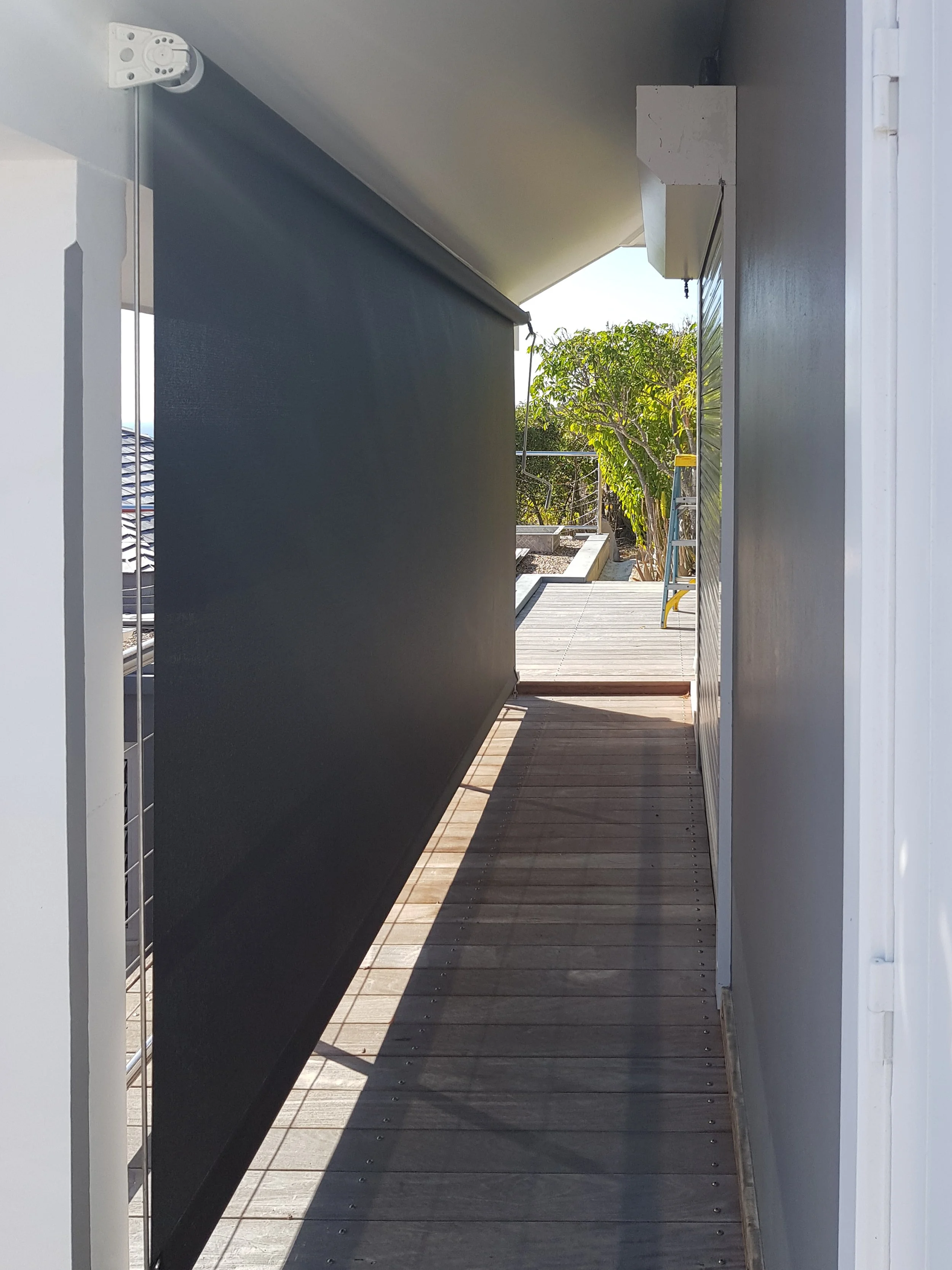 A narrow outdoor walkway with wooden planks, a black roller shade partially pulled down, a ladder in the corner, and trees in the background.