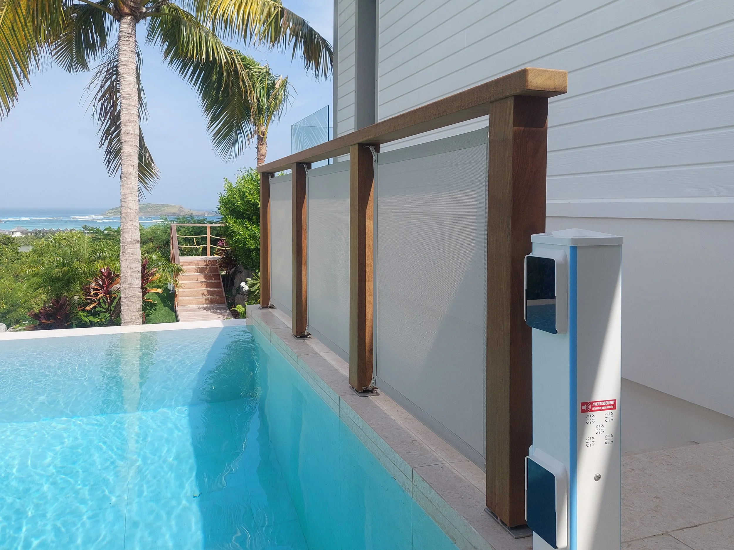 A swimming pool with a view of palm trees, lush greenery, and a distant shoreline with buildings, taken from the edge of the pool area next to a building with white siding. There is a privacy screen and a small white device mounted on the wall.