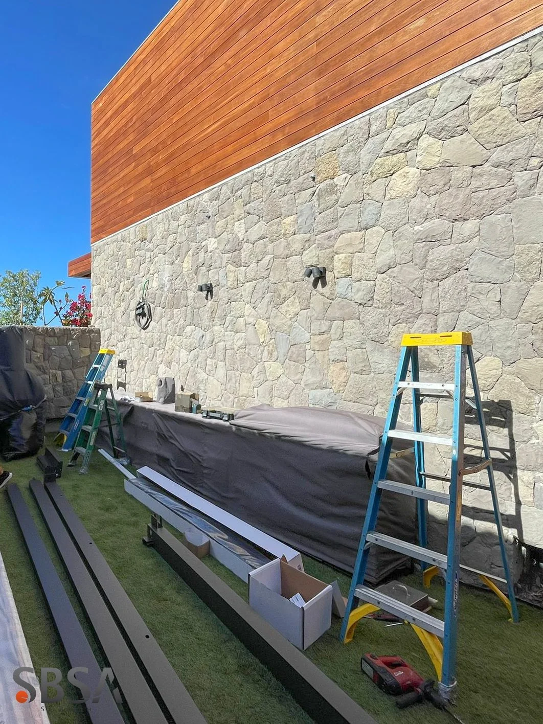 Construction site with a stone wall, ladders, and construction tools, next to a building with wood siding, under a clear blue sky.
