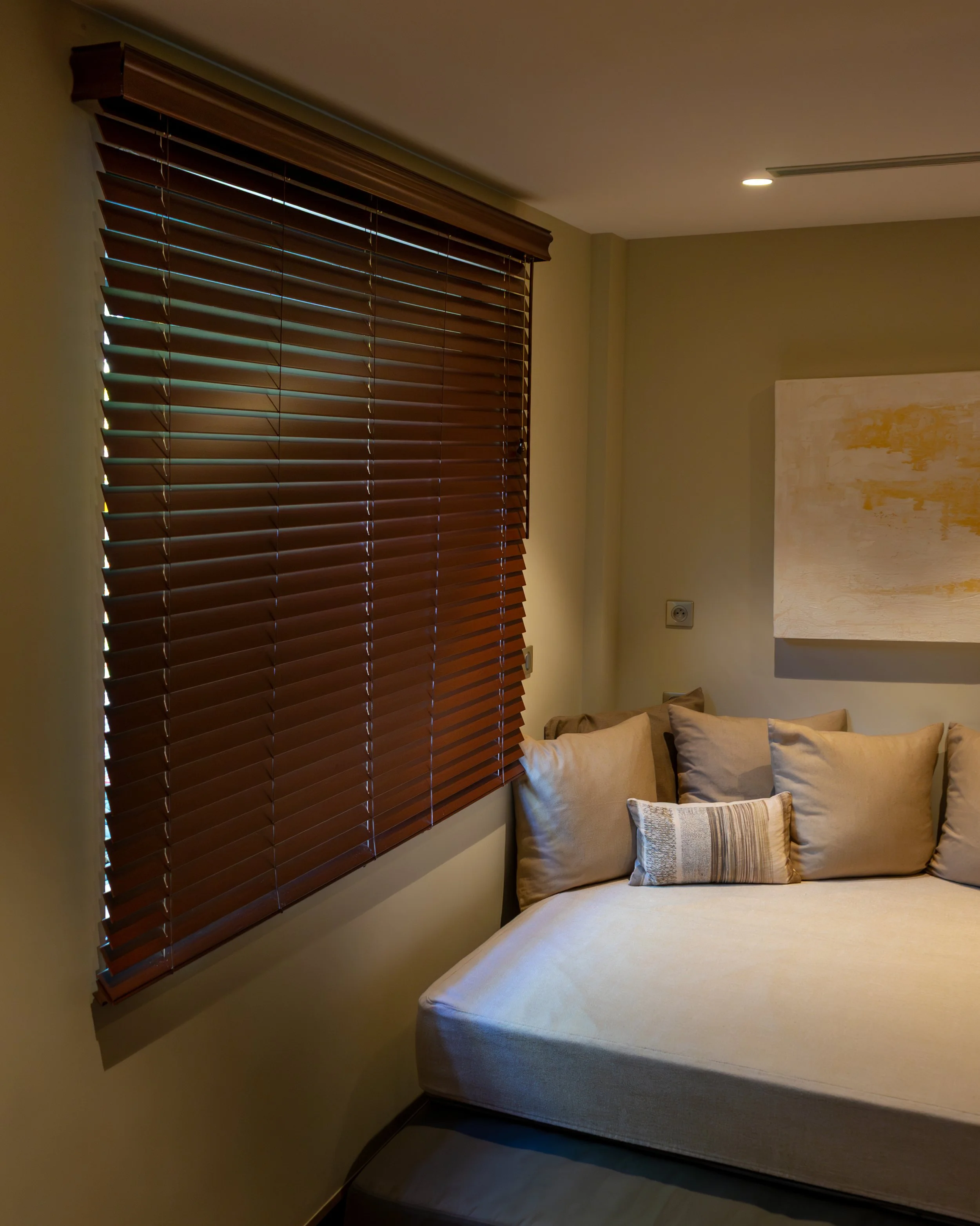 Un coin de chambre avec une fenêtre équipée de stores en bois, un canapé beige agrémenté de plusieurs coussins et une œuvre d’art sur toile accrochée au mur.