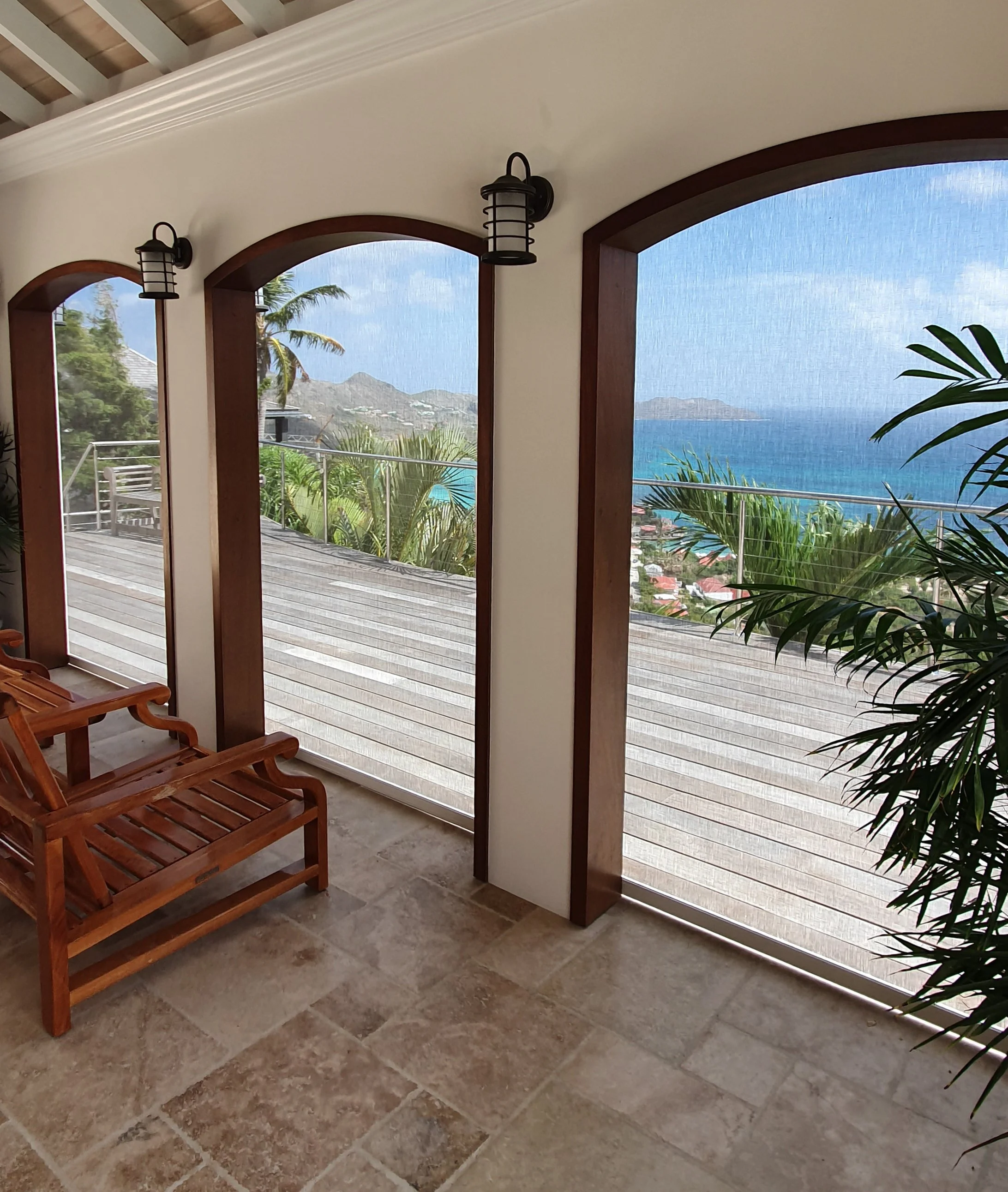 View through arched windows showing outdoor landscape with palm trees, hills, and ocean in the distance, inside a room with beige tiled floor, wooden furniture, and wall-mounted black lantern-style lights.