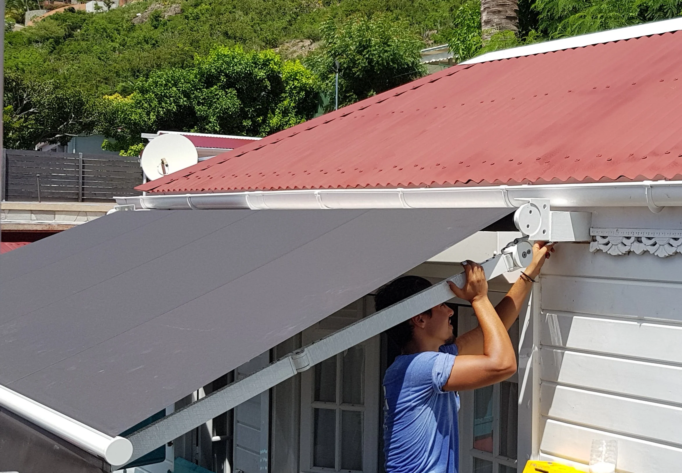 A person working on a roof, handling a metal canopy or awning, with a house, trees, and a satellite dish in the background.