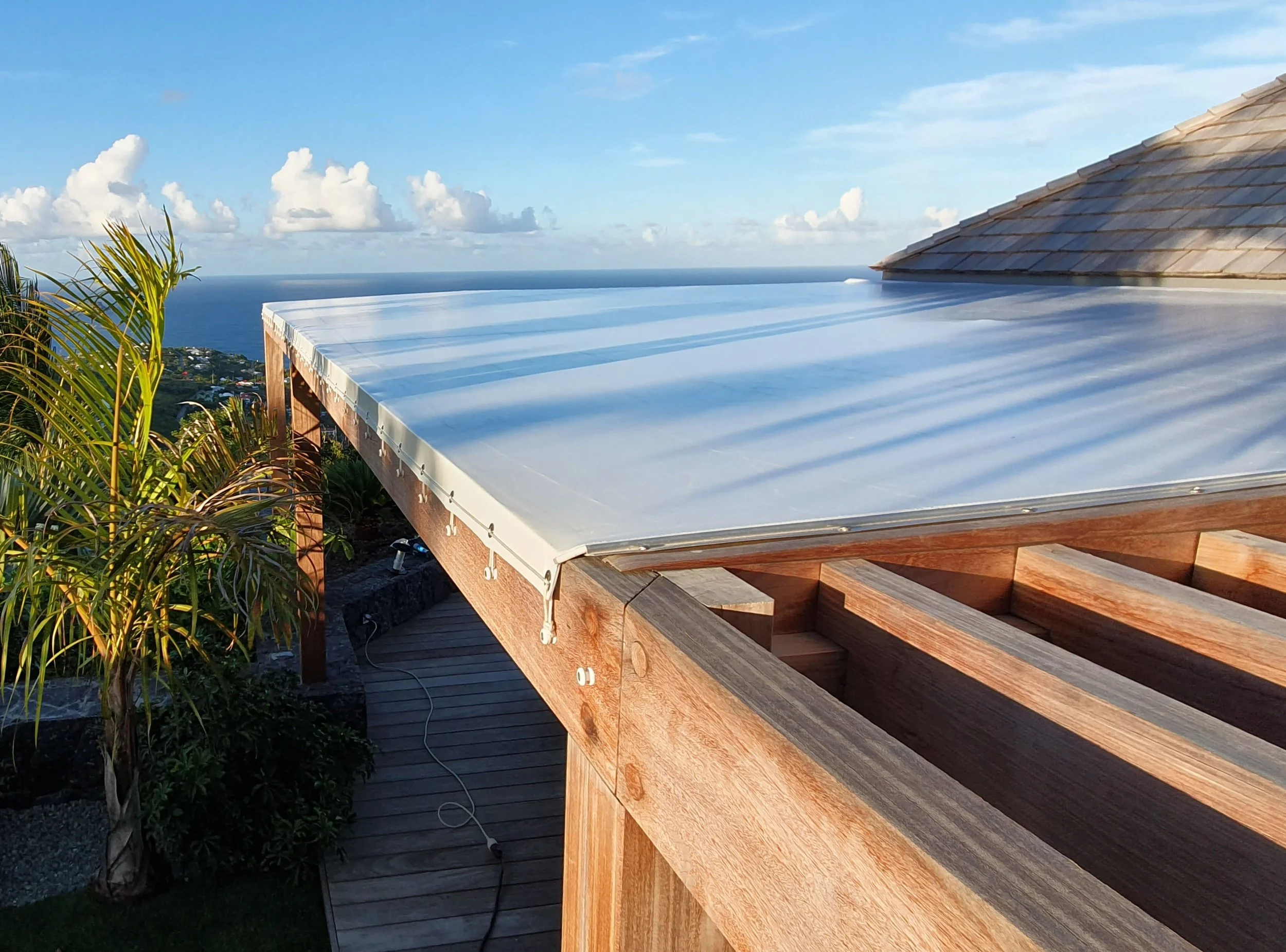 A rooftop with a stainless steel or aluminum covering, set against a backdrop of a blue ocean and sky with scattered white clouds, and some greenery on the side.
