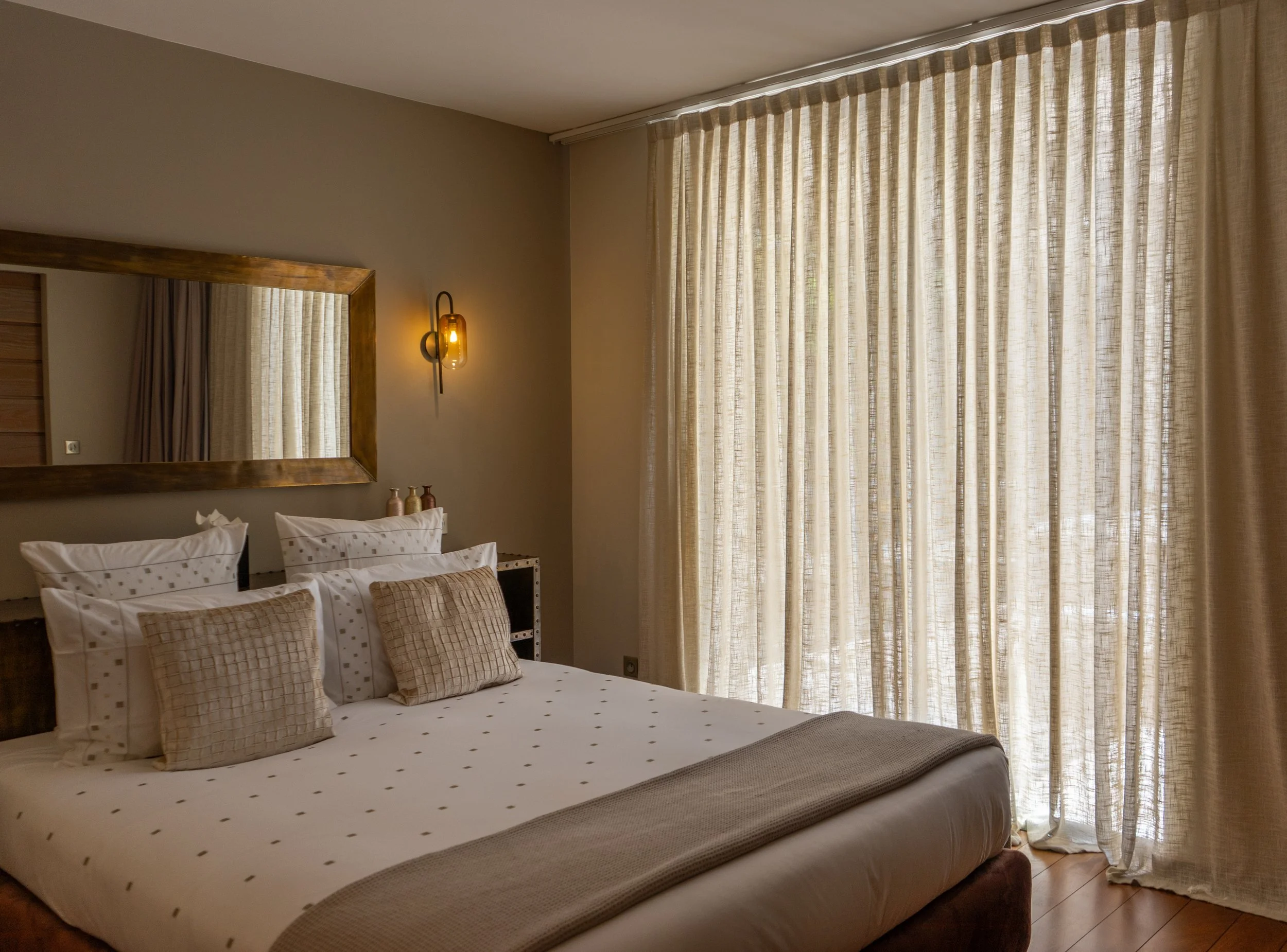 Chambre chaleureuse avec un grand lit, une literie beige et plusieurs oreillers aux motifs assortis. Un miroir encadré de bois est suspendu au-dessus du lit, tandis qu’une applique murale diffuse un éclairage doux et chaleureux. De doux rideaux beige