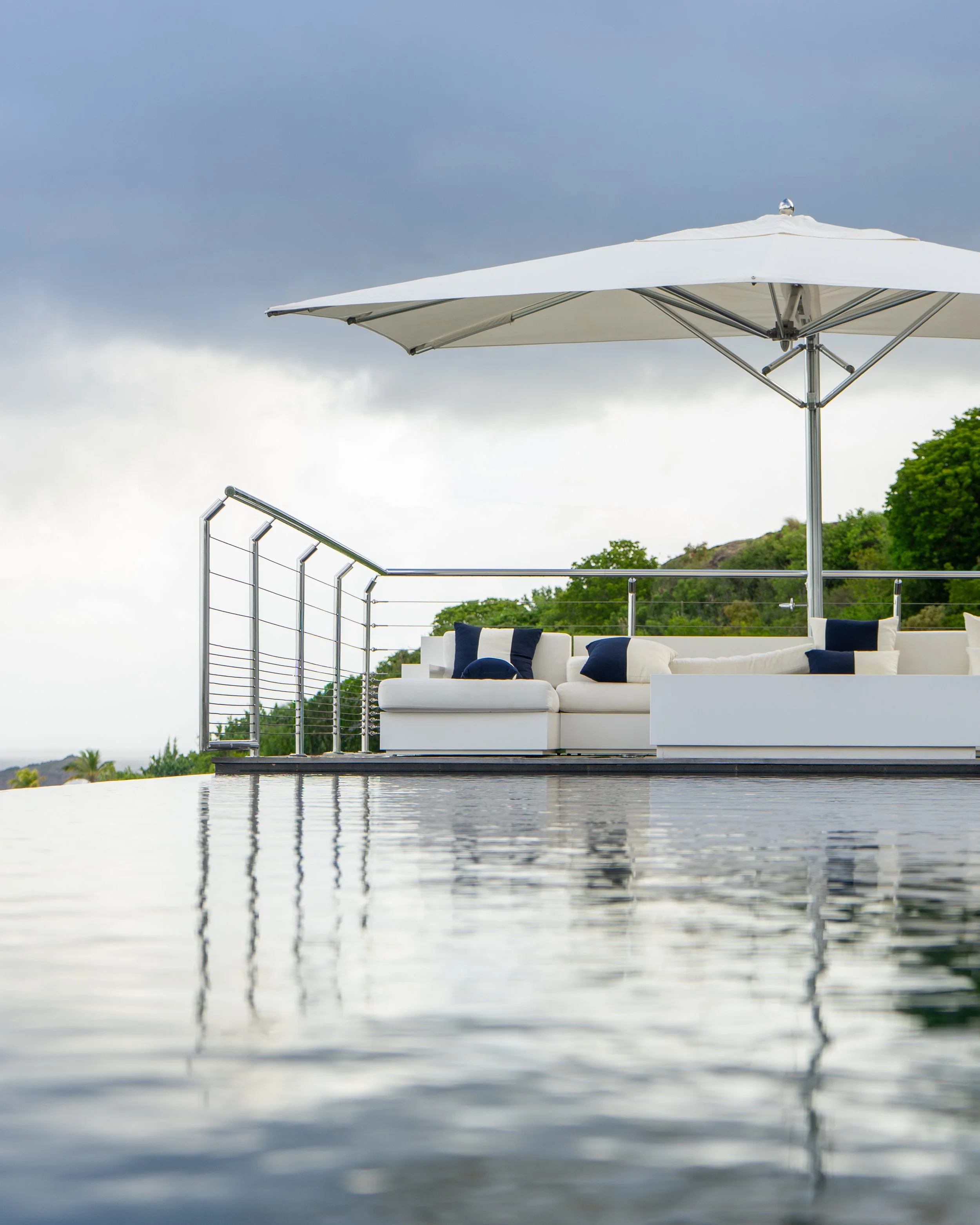 Outdoor scene with a large white patio umbrella, white outdoor seating adorned with navy and white pillows, a metal railing, a cloudy sky, and green trees in the background.