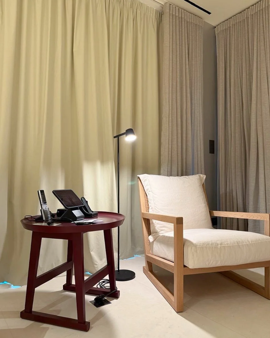 A room with beige curtains, a wooden armchair with cream cushions, a round red side table holding a cordless phone, a tablet, and a lamp, and a floor lamp next to the table.