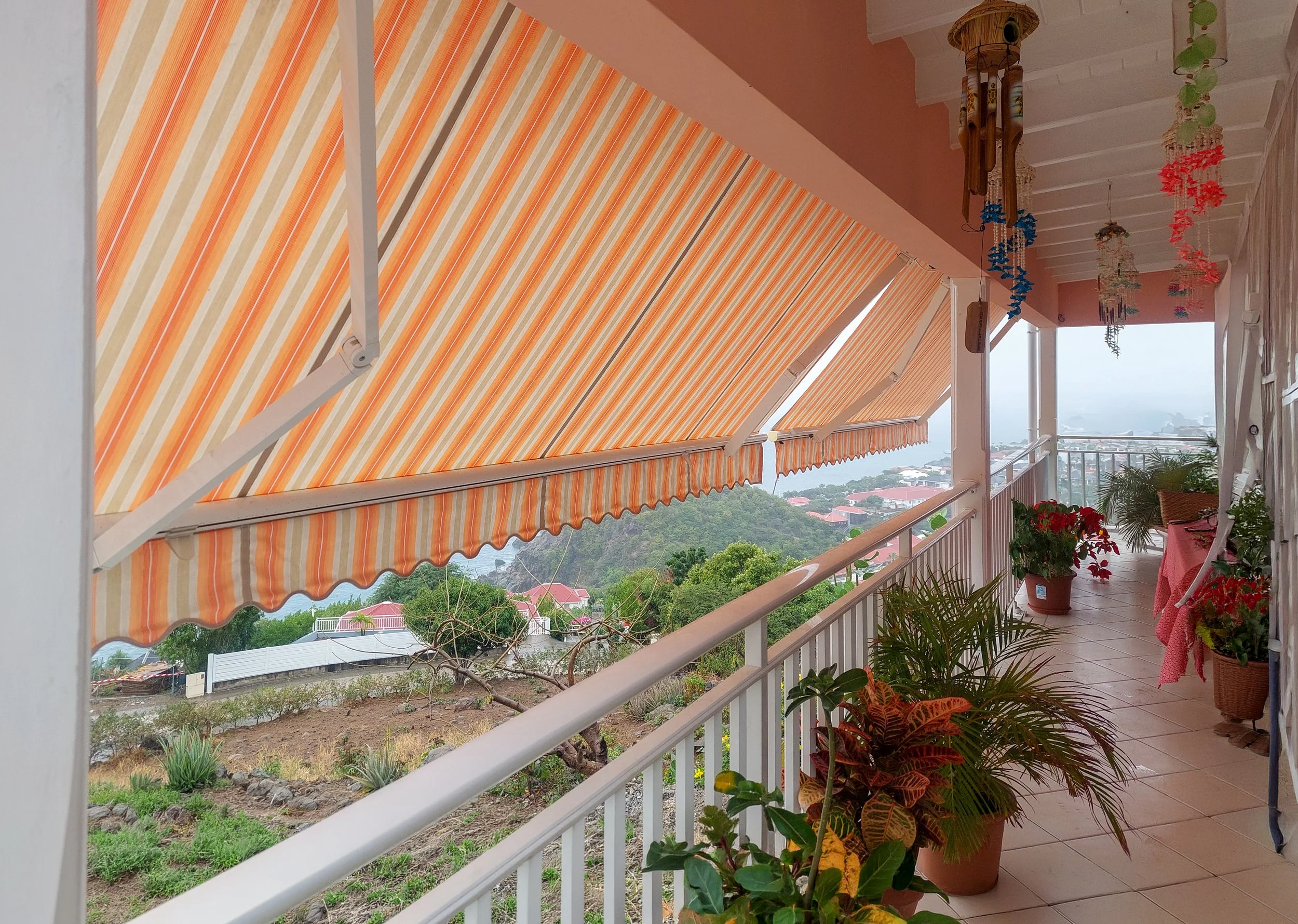 A balcony with potted plants, colorful hanging decorations, and an orange striped retractable awning overlooking a hilly area with houses and greenery.