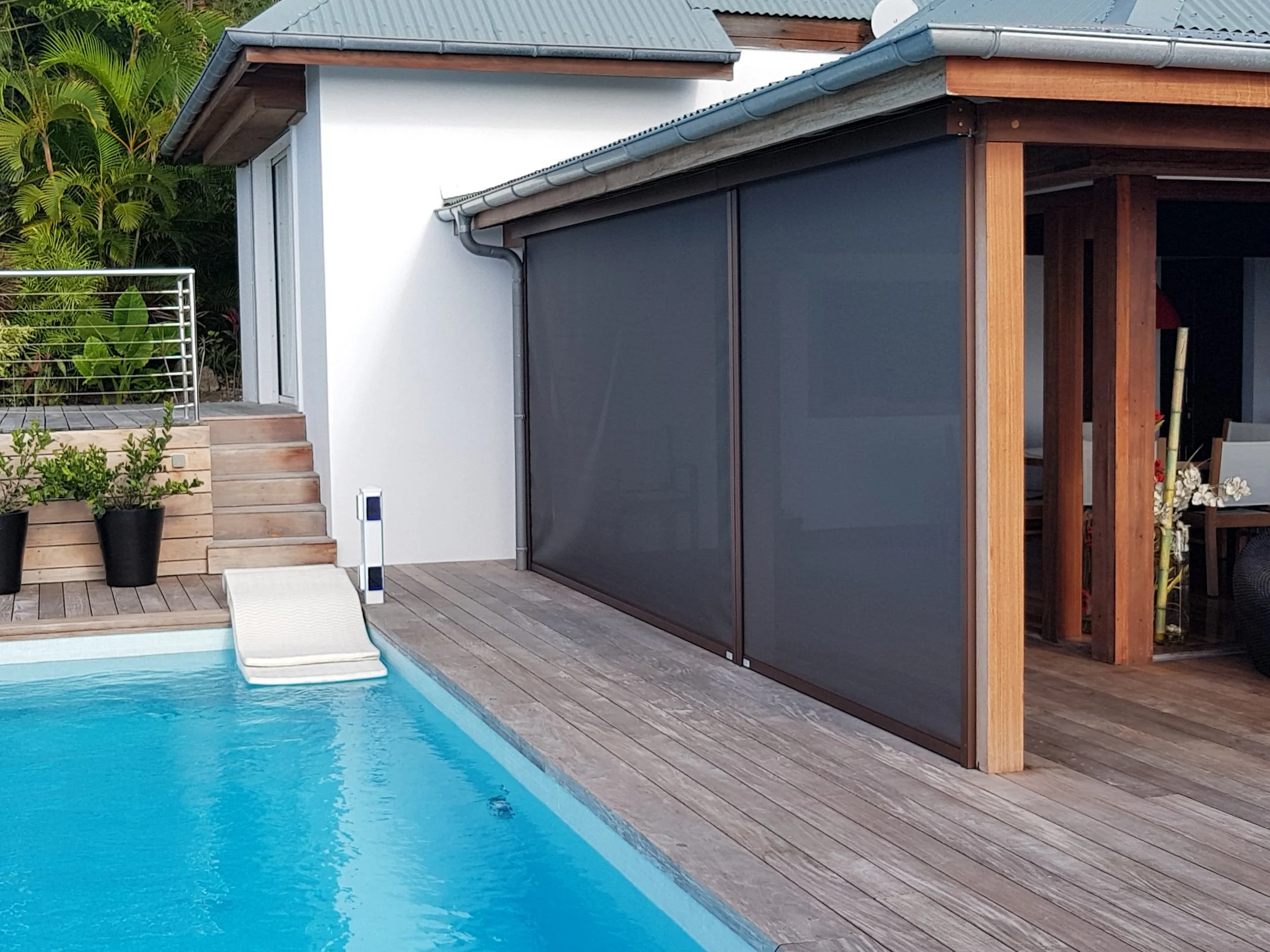 A modern house with wooden accents and dark grey shading, next to a swimming pool with wooden decking, lush green plants, and outdoor furniture.