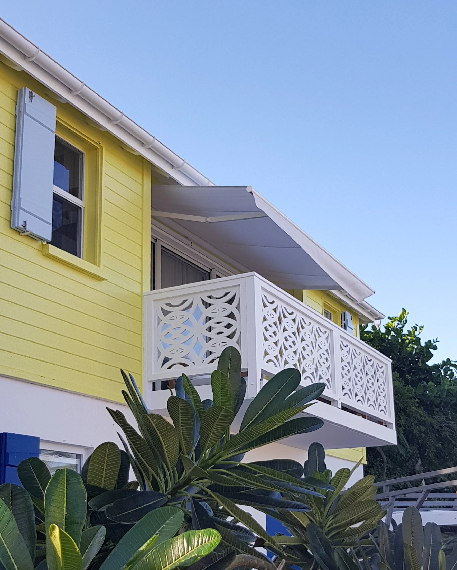 Yellow house with white balcony railing and blue shutters, surrounded by lush green leaves, under a clear blue sky.