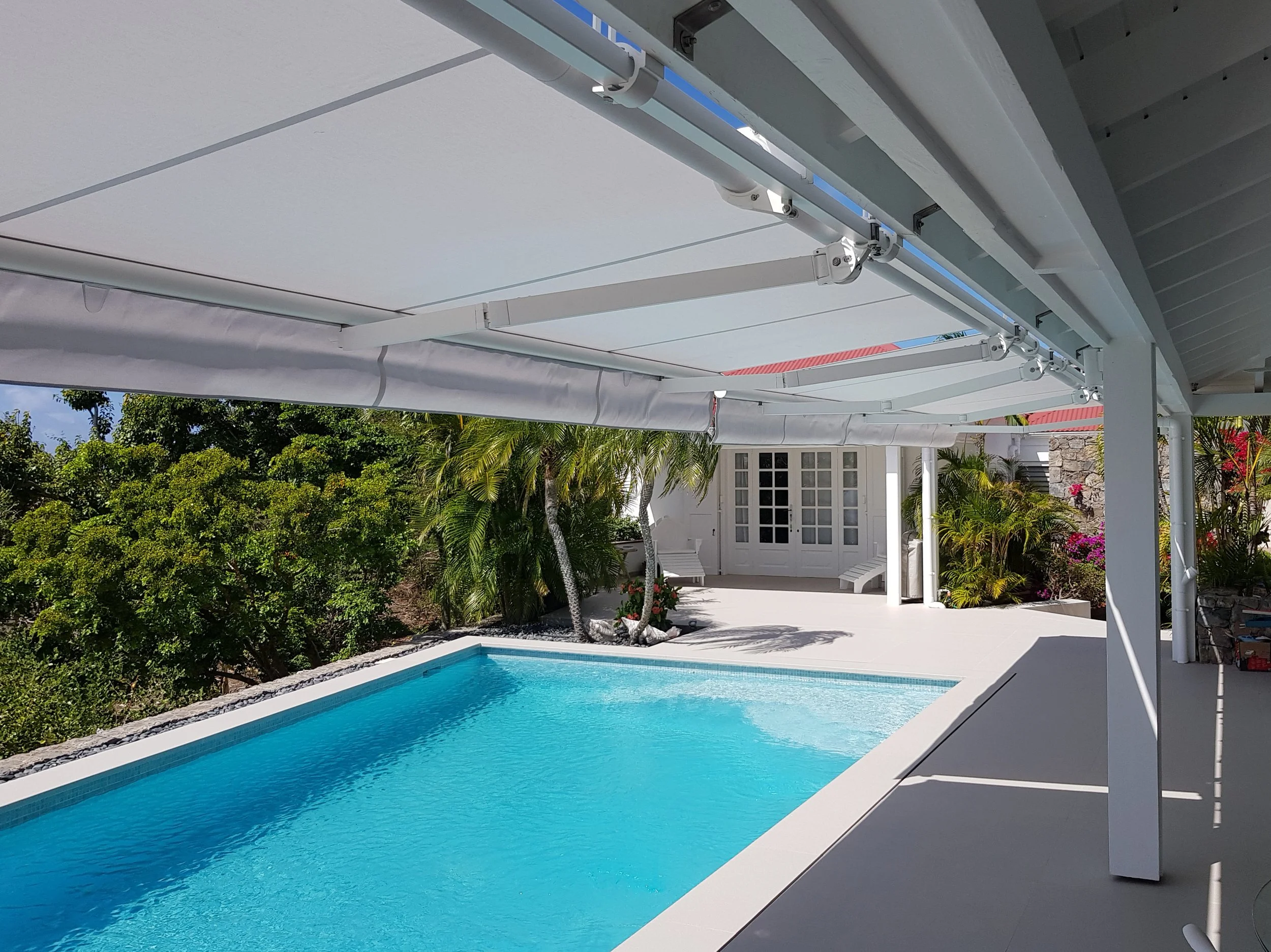 A backyard swimming pool area with clear blue water, white pool deck, and lush green plants and trees surrounding it. There are white patio chairs and a shaded area with a white pergola or canopy.