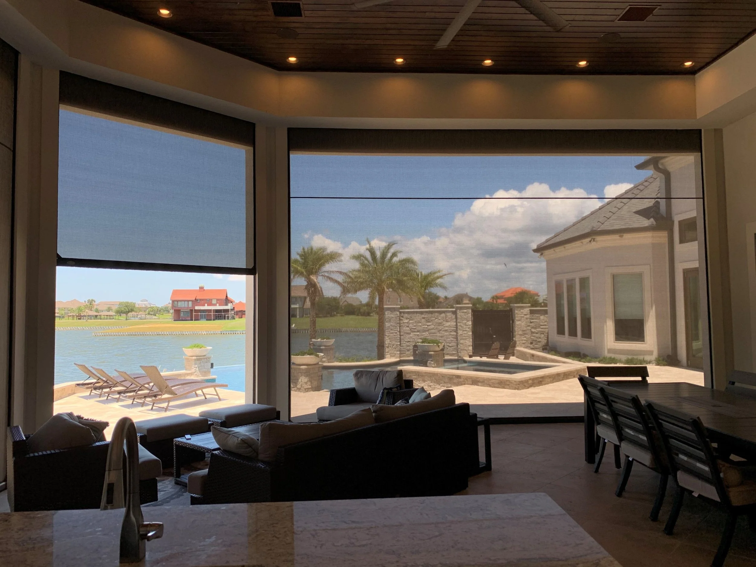 View from inside a house looking out through a large screened-in porch to a backyard with palm trees, a lake, and houses in the distance. The porch has outdoor furniture including a sofa, chairs, and lounge chairs.