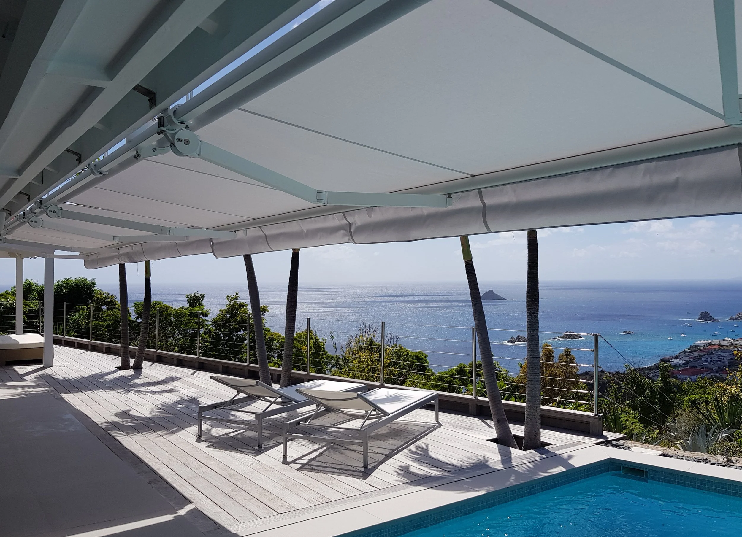 View of a balcony overlooking the ocean with palm trees, a swimming pool, white lounge chairs, and an awning under a partly cloudy sky.