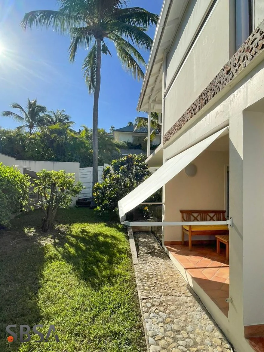 View of a backyard with lush green grass, small bushes, and tall palm trees under a bright blue sky. Part of a white house with a patio featuring a wooden bench and a white awning is visible on the right side.