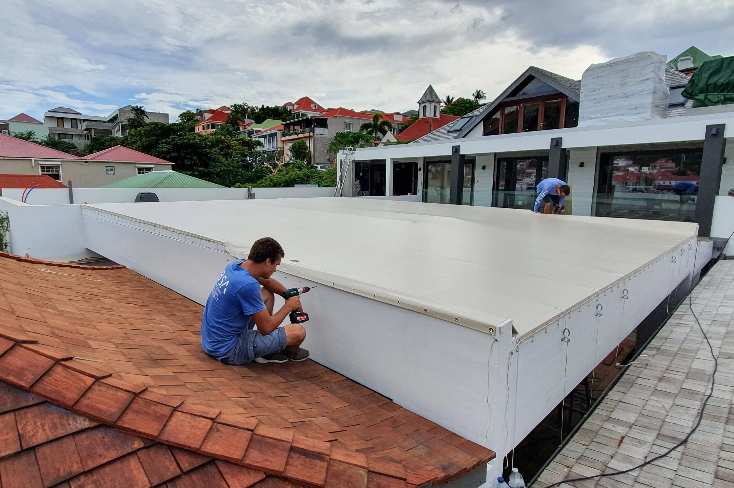 Two workers installing a large white retractable awning on a house's patio. One worker is kneeling and using a drill, while the other is standing and working on the awning's mechanisms. The background shows multicolored residential buildings on a hillside with a cloudy sky.