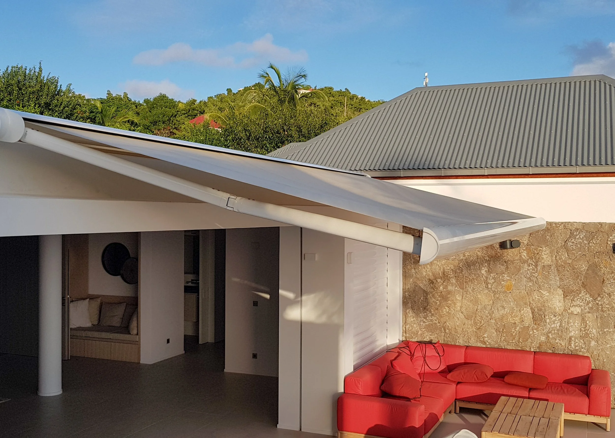 A view from inside a house showing a living area with a red sectional sofa and a wooden coffee table, kitchen cabinets, and a open doorway leading to another room. Outside the window, there is greenery and a house with a metal roof, and in the backgr