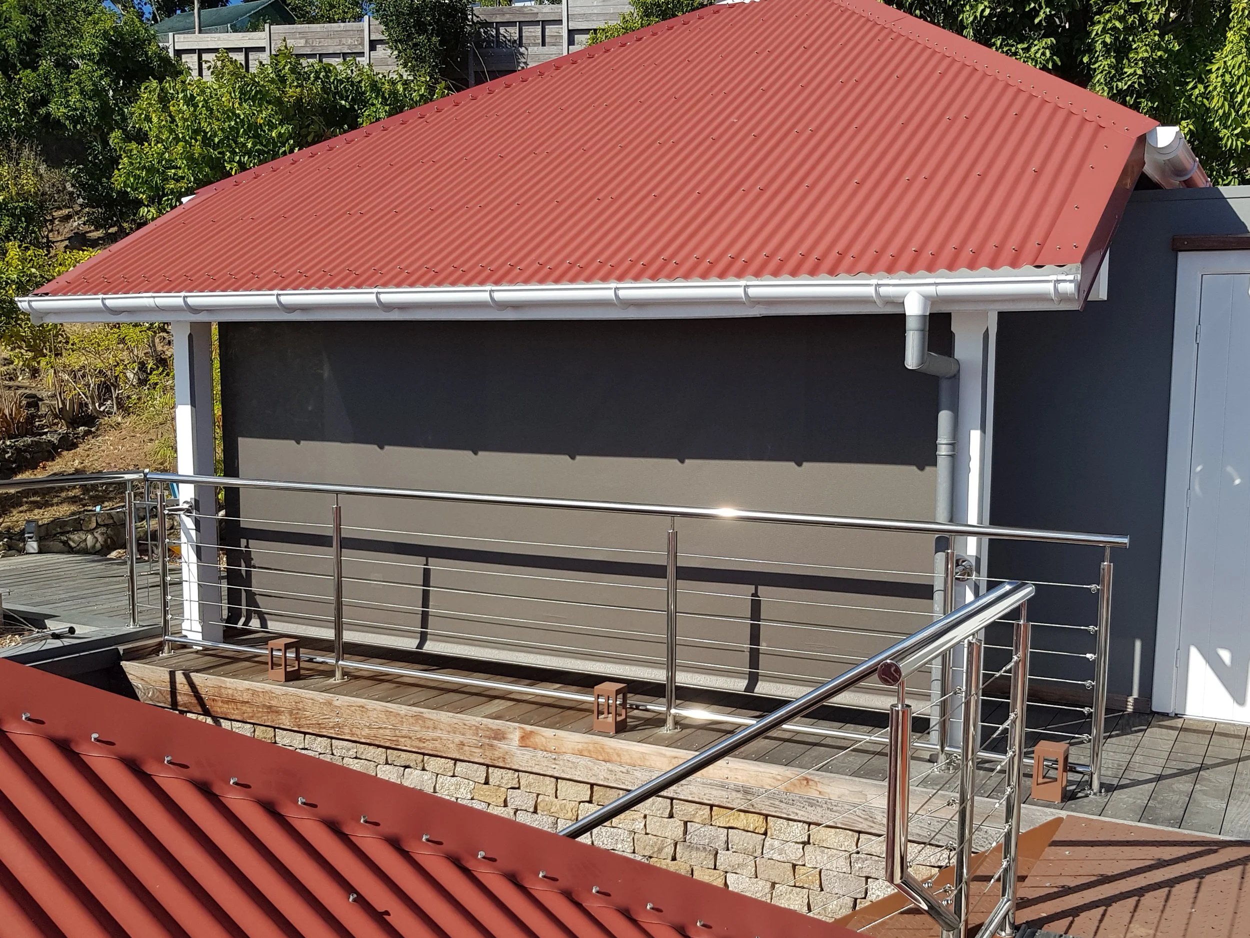 Red metal roof with a white gutter over a gray building, a balcony with a metal railing, and a lush green garden in the background.