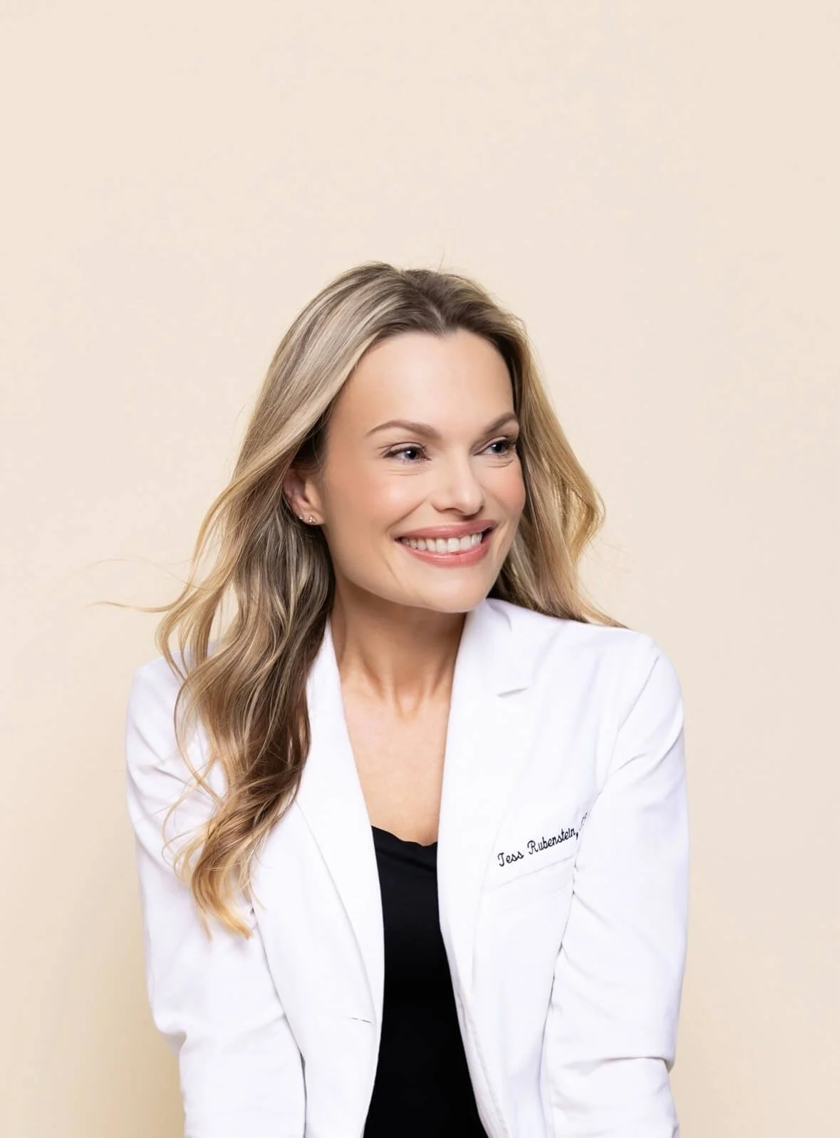 A smiling woman with long, wavy blonde hair wearing a white coat and black top, looking to her right, posing against a plain beige background.