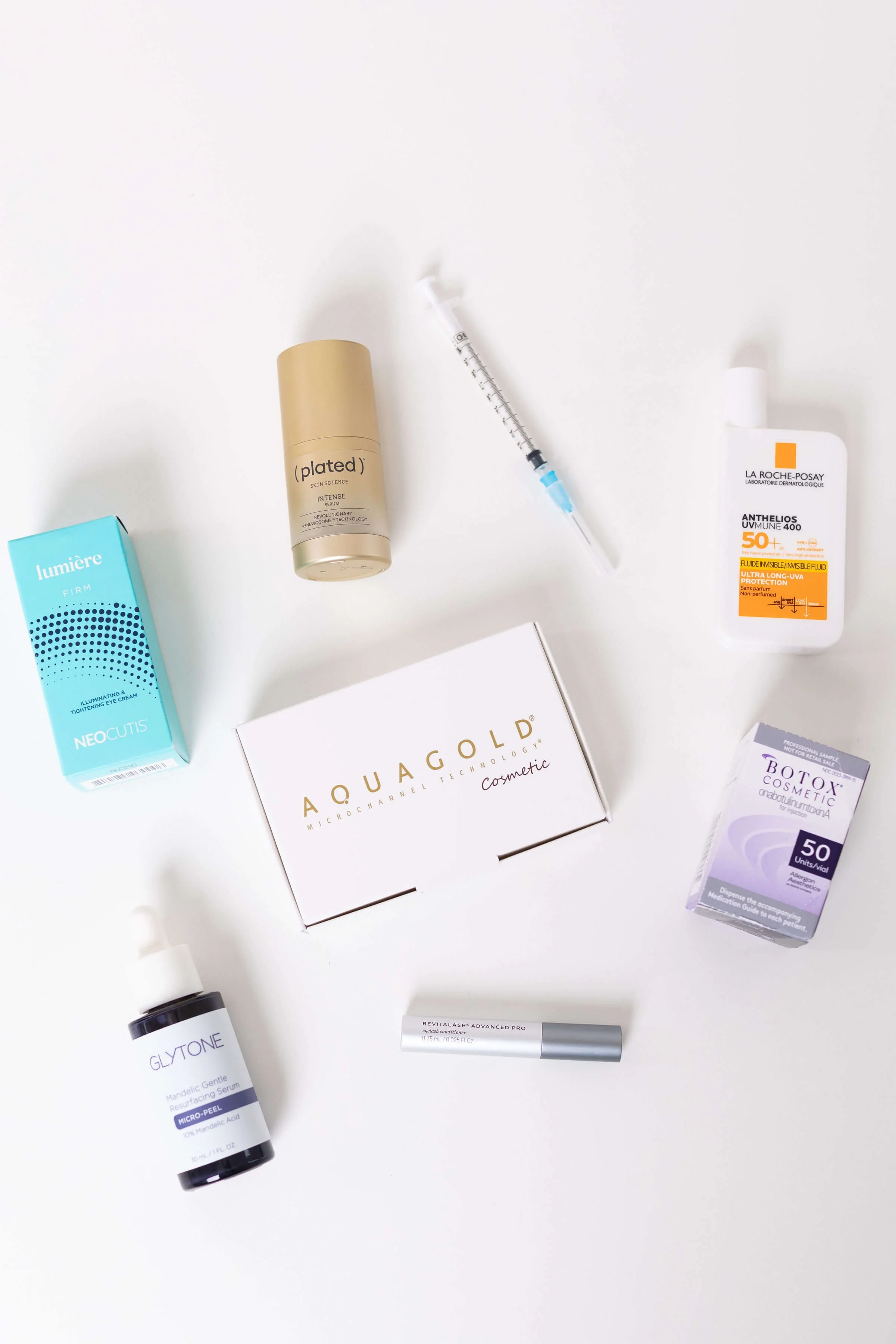 Collection of skincare and cosmetic products on a white surface, including a blue Lumiere firm illuminating eye cream, a gold Plated skin science serum, a white La Roche-Posay sunscreen, a white box labeled Aquagold, a purple Botox cosmetic box, a gray Revitalash serum, and a black Glytone serum.