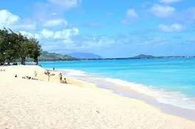 Sandy beach with a few people, trees on the left, clear blue sky, and turquoise ocean with distant islands.