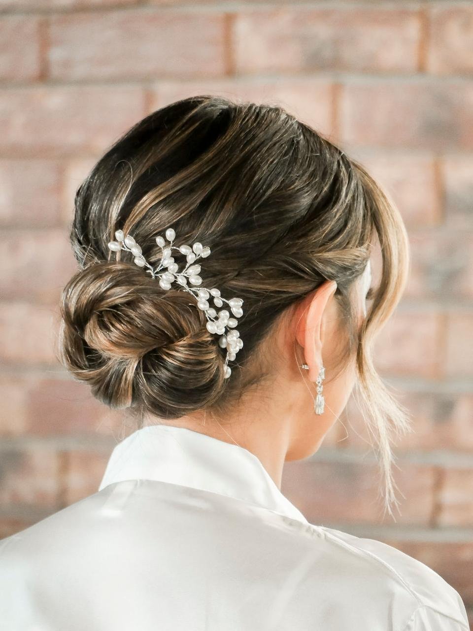 Peinado de novia con recogido trenzado adornado con broche de perlas, vista desde atrás, con pendientes elegantes y blusa blanca, fondo de ladrillo.
