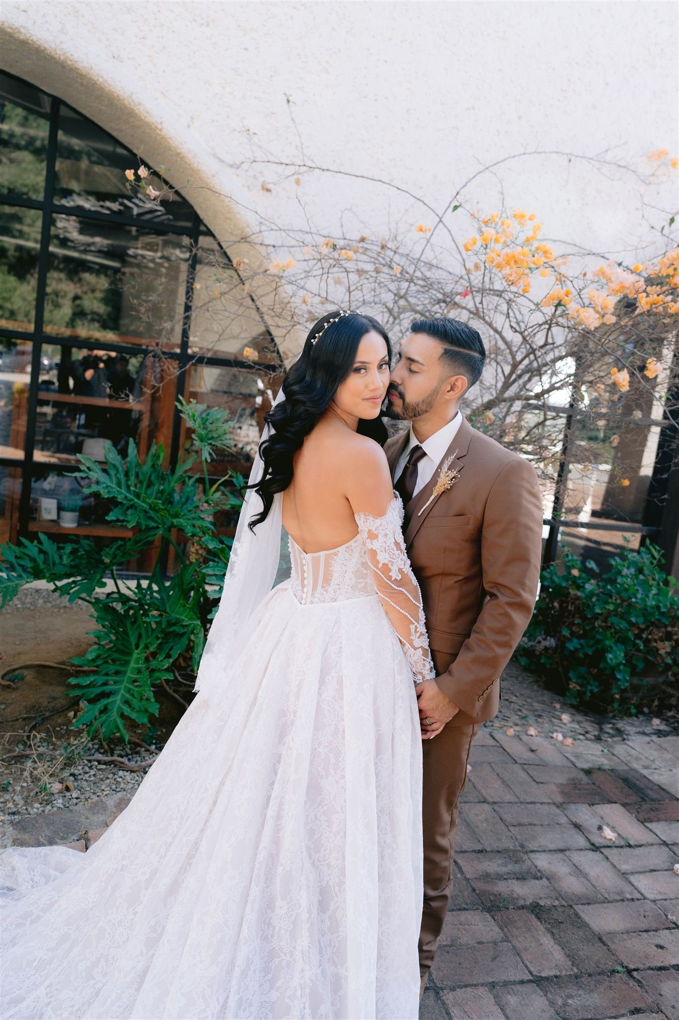 Una pareja en su boda, la mujer lleva un vestido blanco de encaje y el hombre un traje marrón, en un espacio con plantas y ramas con flores amarillas en el fondo.